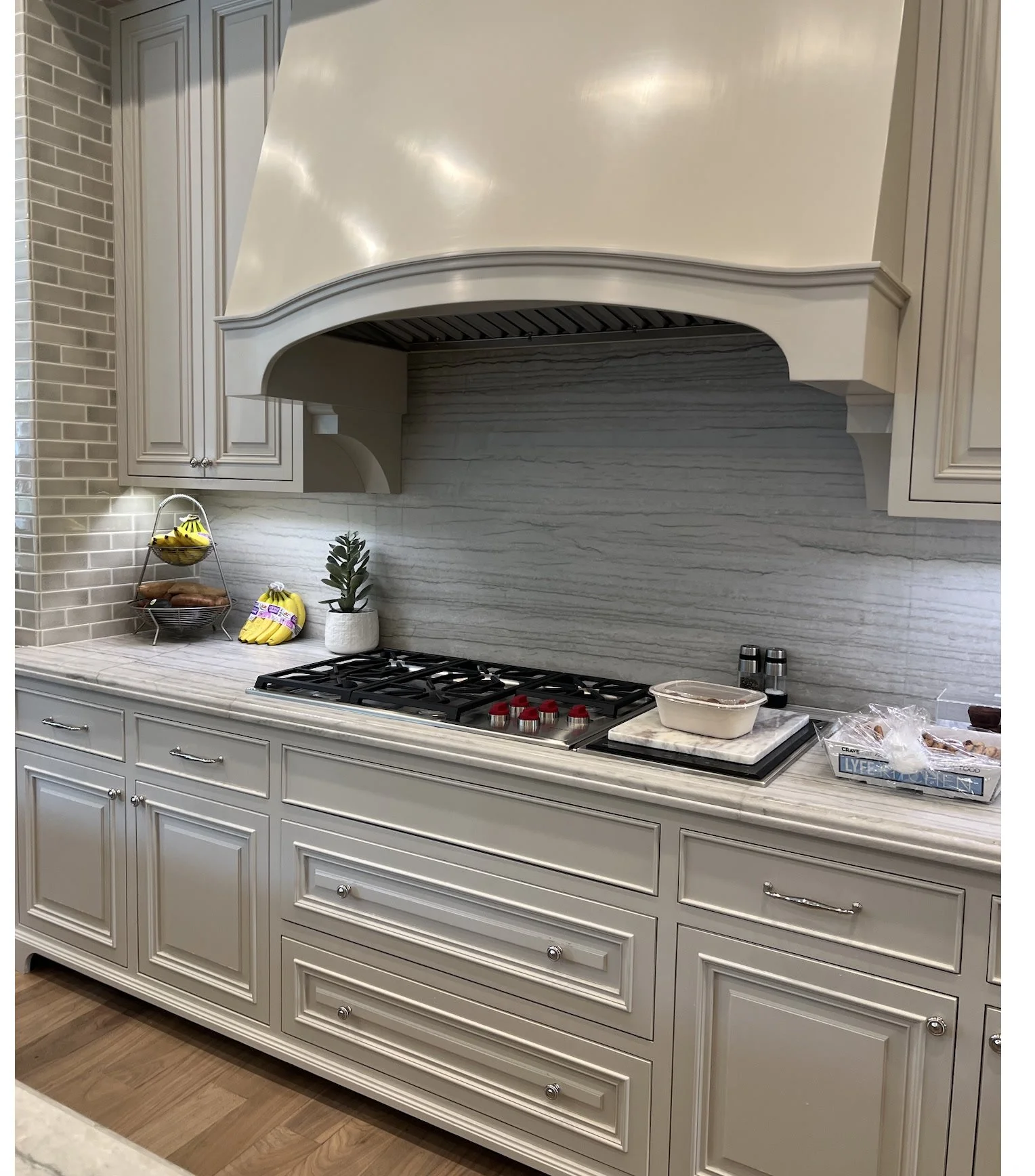 The kitchen before renovation: ornate gray-sage cabinetry, mantel hood, chrome hardware, subway tile, and a gas rangetop.
