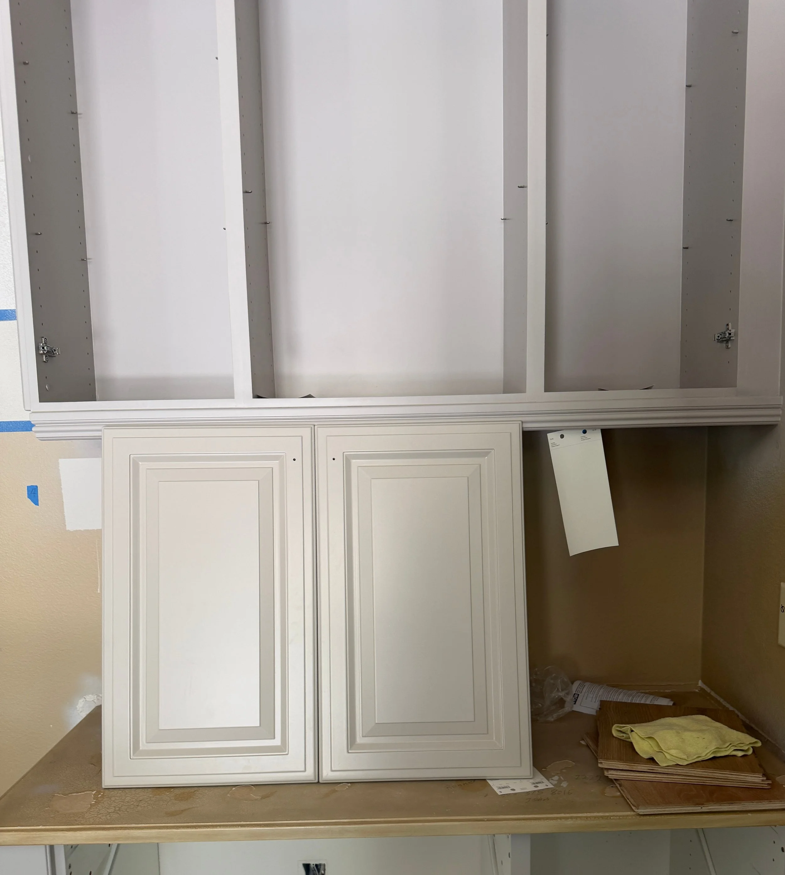 Kitchen job site with cream cabinet doors leaning on the counter, paint swatches, and unfinished cabinet boxes — testing color in real light.