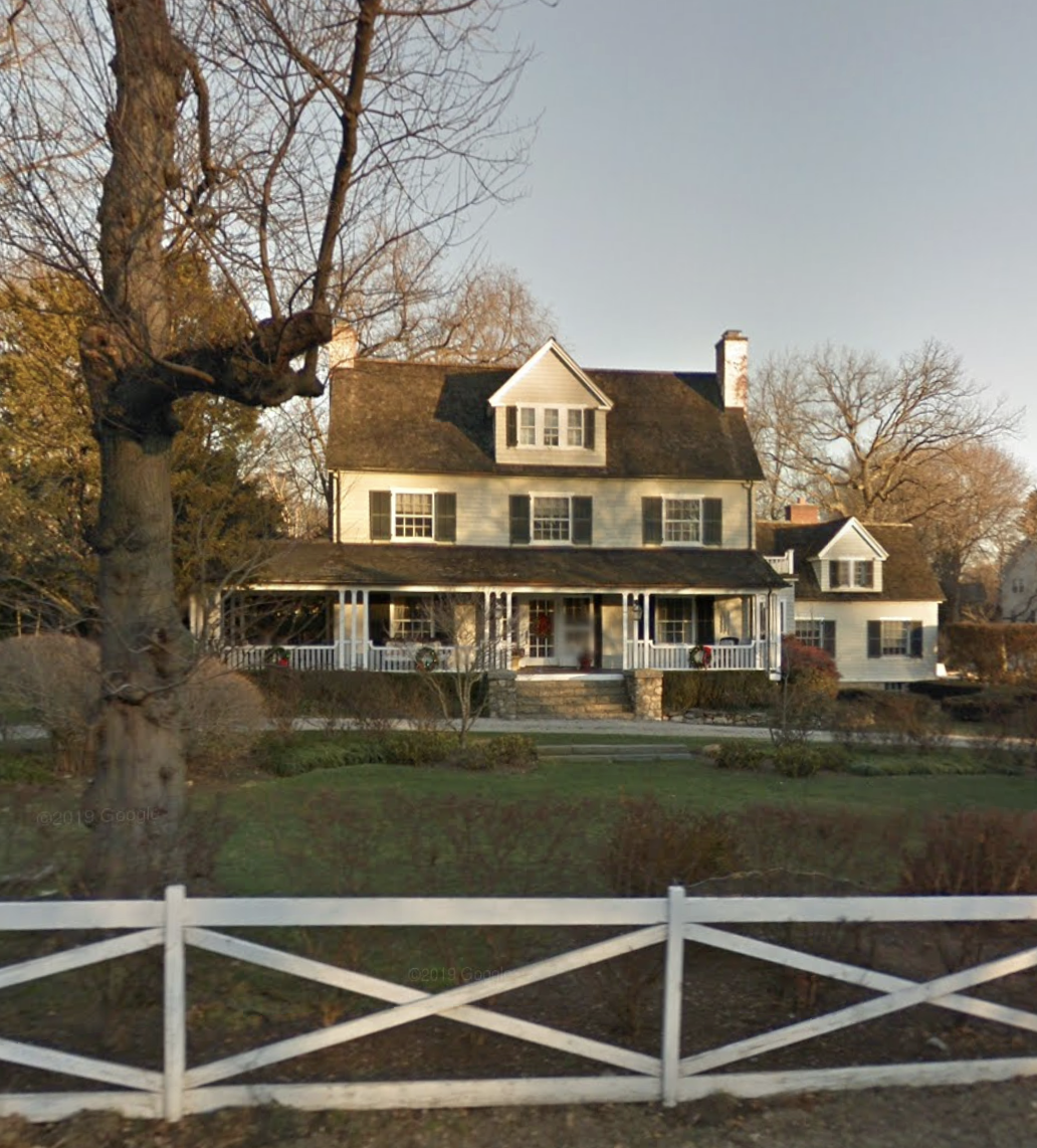 White colonial farmhouse with a wide front porch behind a white split-rail fence and a large bare tree