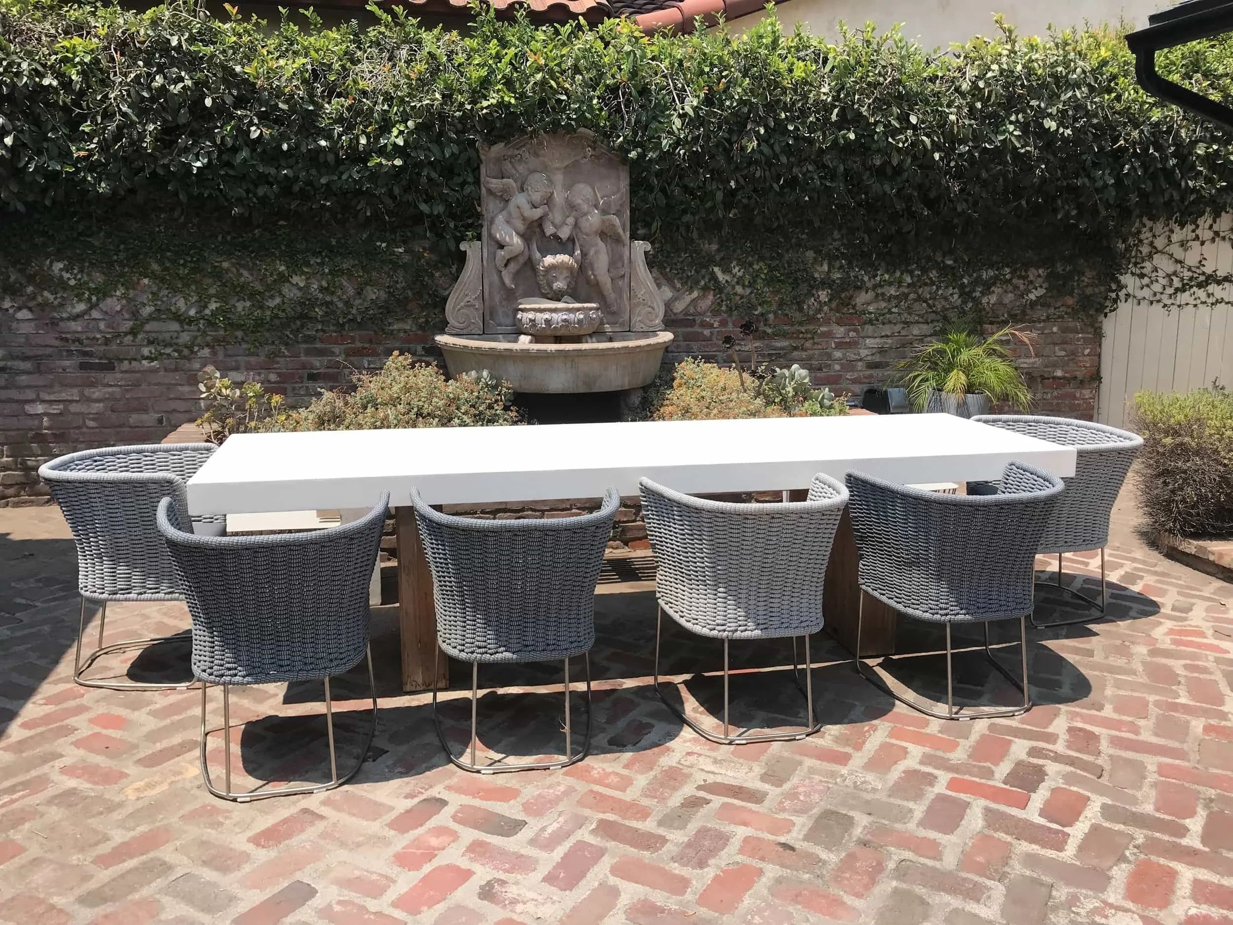 Outdoor patio dining area with a long white table, six woven gray chairs, a brick floor, a brick wall with a water fountain featuring cherub statues, and potted plants.