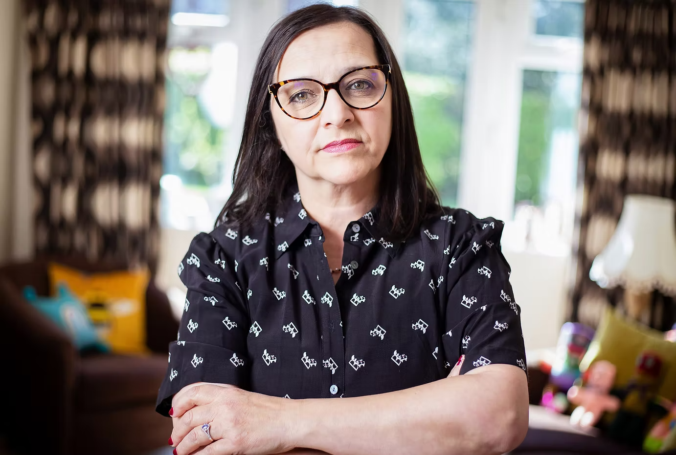 A woman with dark hair, glasses, and a black shirt with white patterns, standing indoors with arms crossed in front of a window with curtains.
