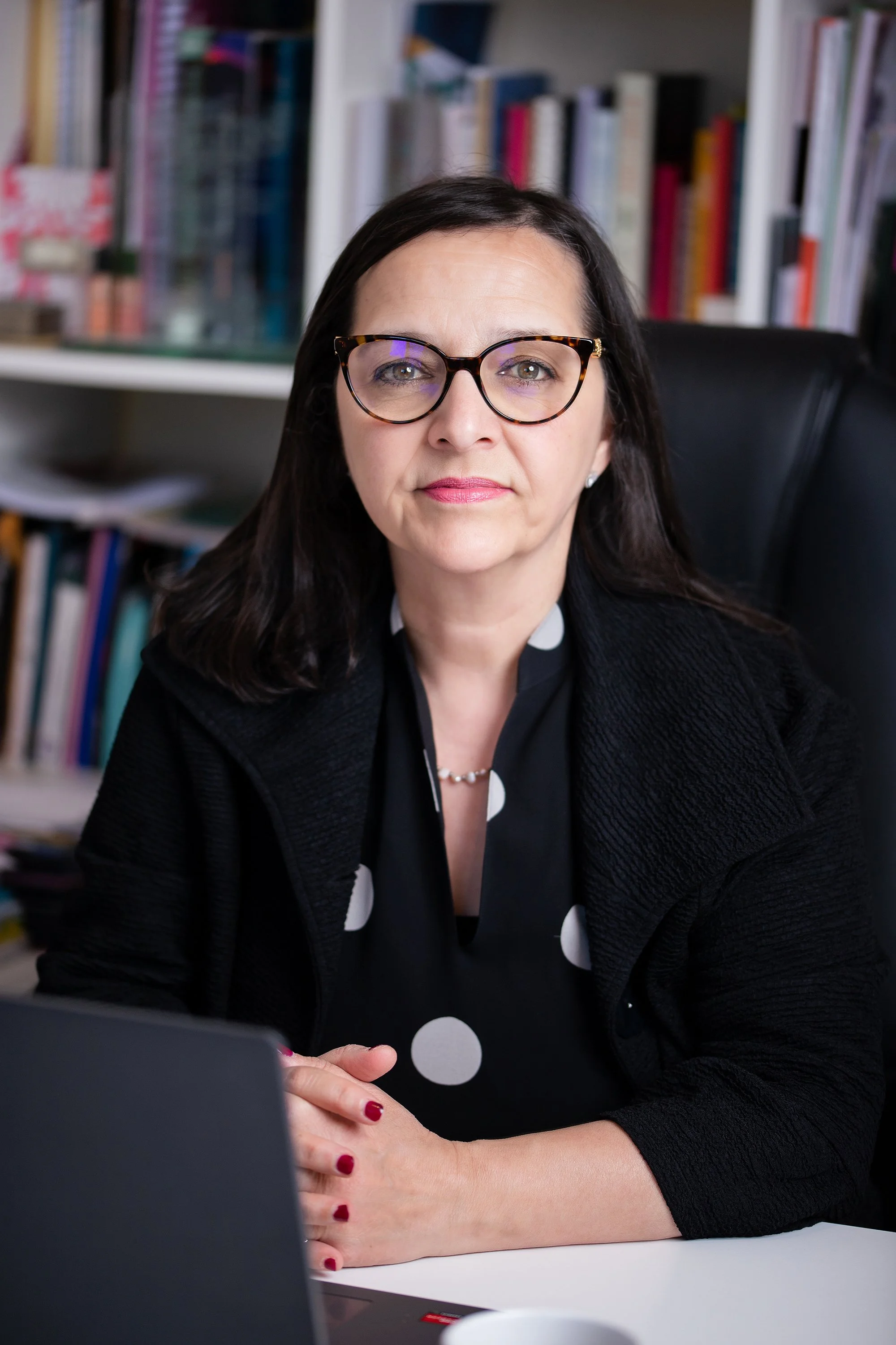 A professional woman with dark hair, glasses, and a black blazer sits at a desk in an office with shelves of books behind her.