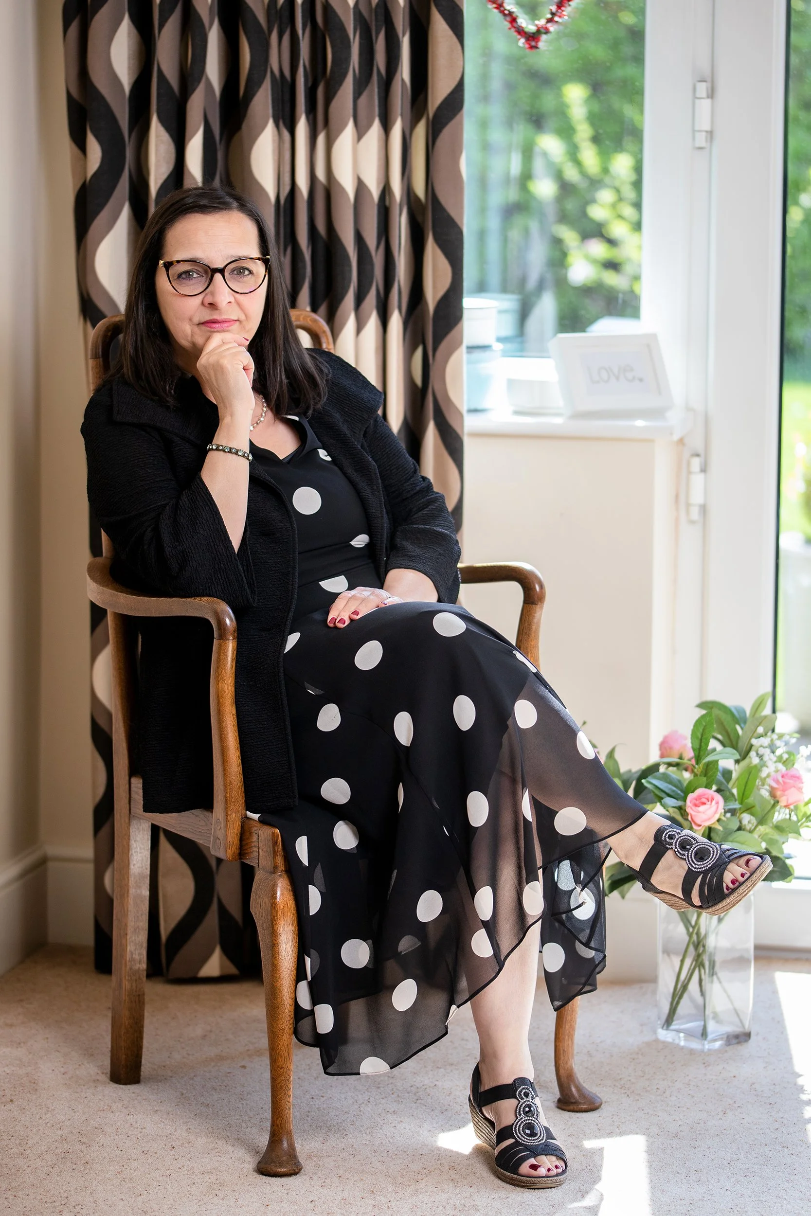 A woman sitting in a wooden chair indoors near a window, wearing a black dress with white polka dots, a black cardigan, and black sandals with circular designs. She has glasses, dark hair, and is posing with her chin resting on her hand, looking directly at the camera.