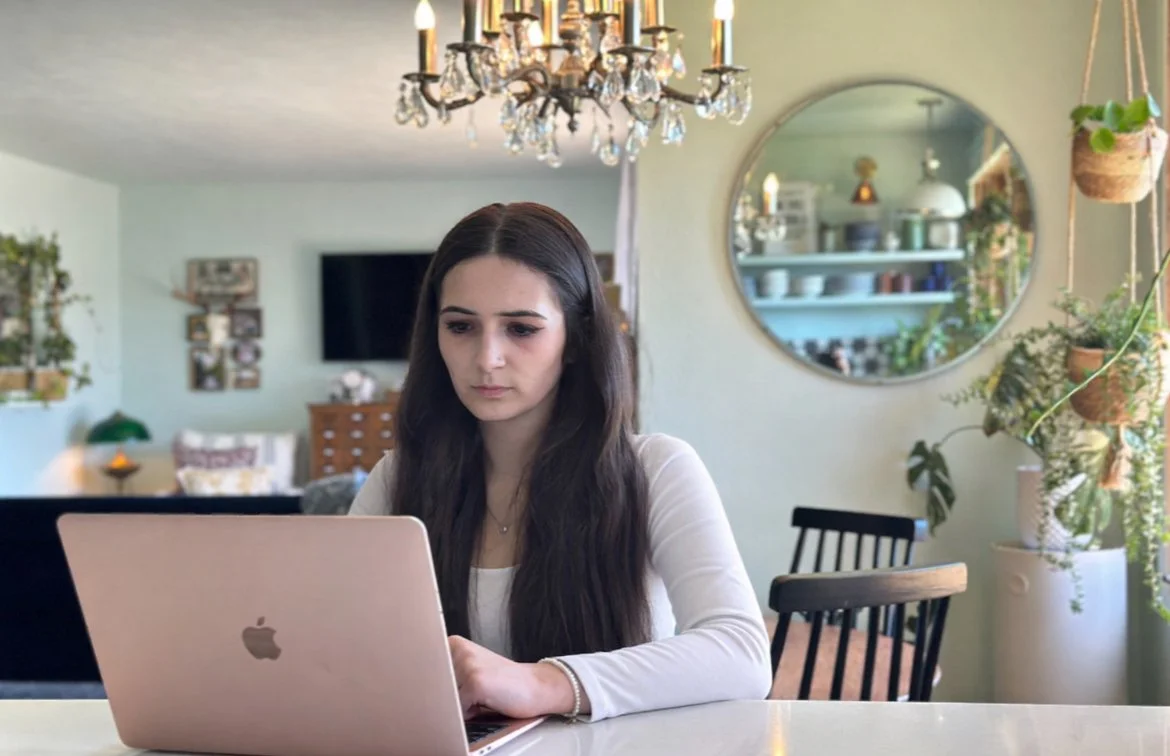 Woman with long dark hair working on a silver MacBook at a dining table in a well-lit, decorated living room with plants, a chandelier, and a round mirror.