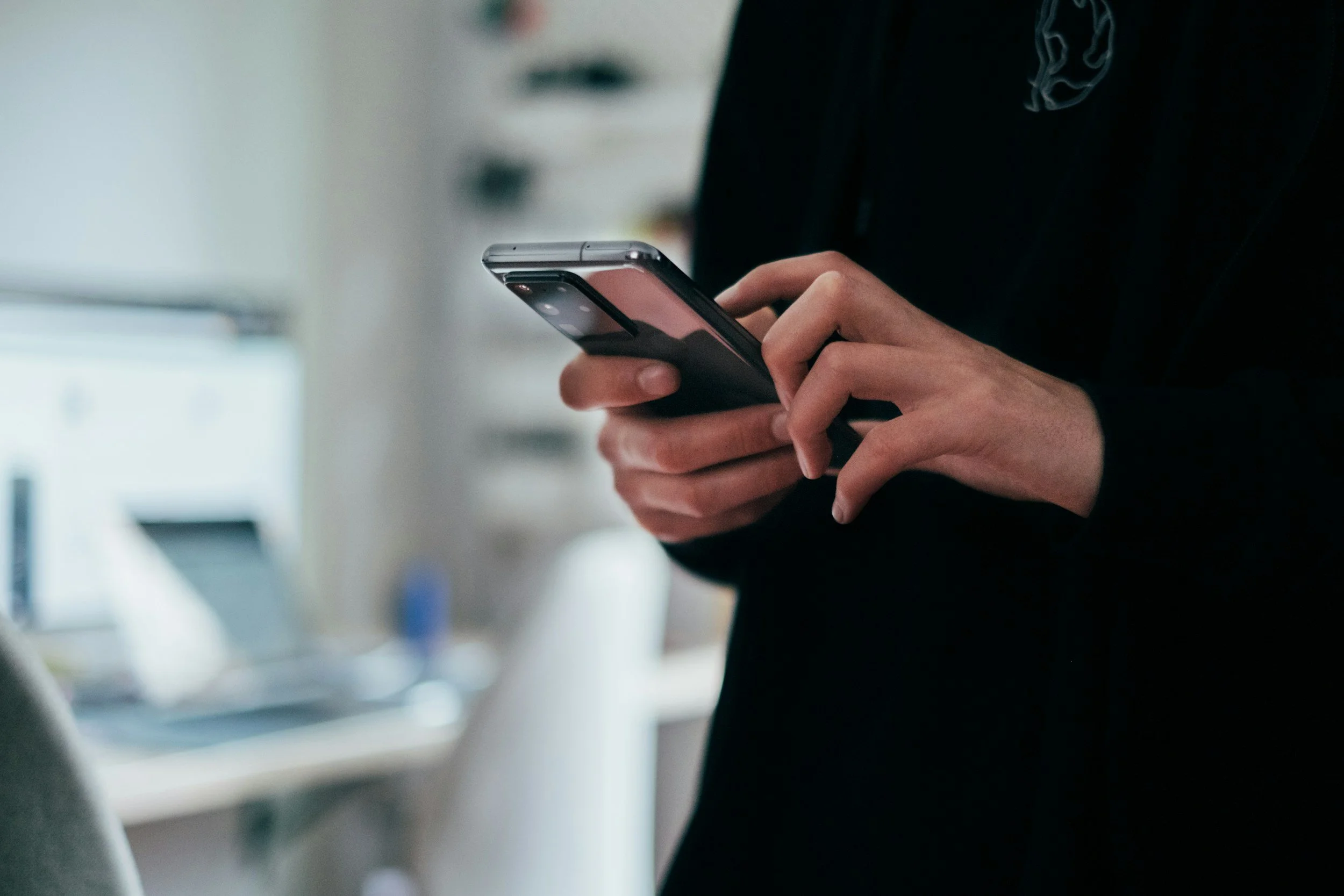 Person holding a smartphone in a room with a computer and shelves in the background.