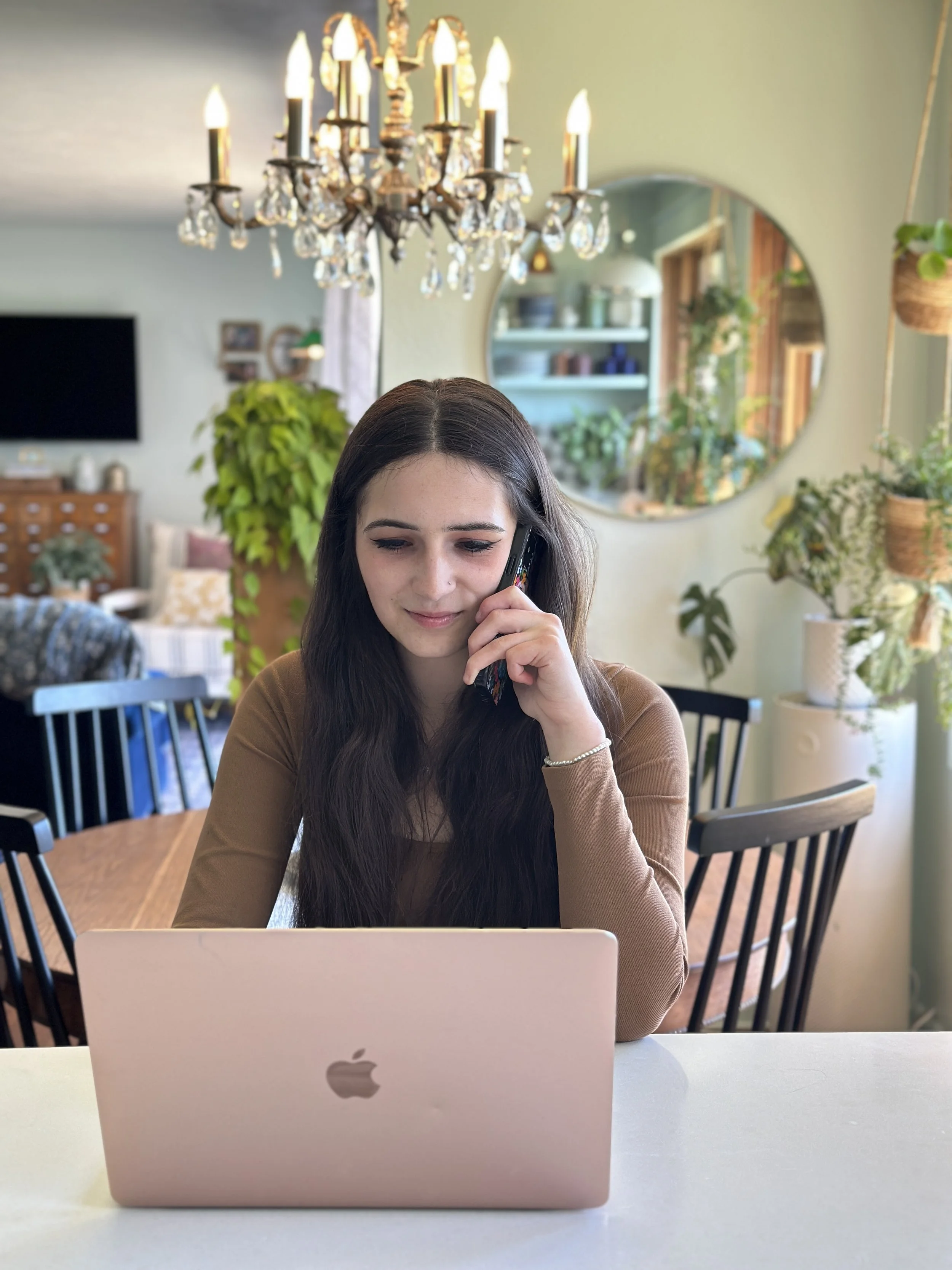 Young woman with long dark hair talking on a cell phone while sitting at a table with a pink MacBook in a well-lit room with plants and a chandelier.