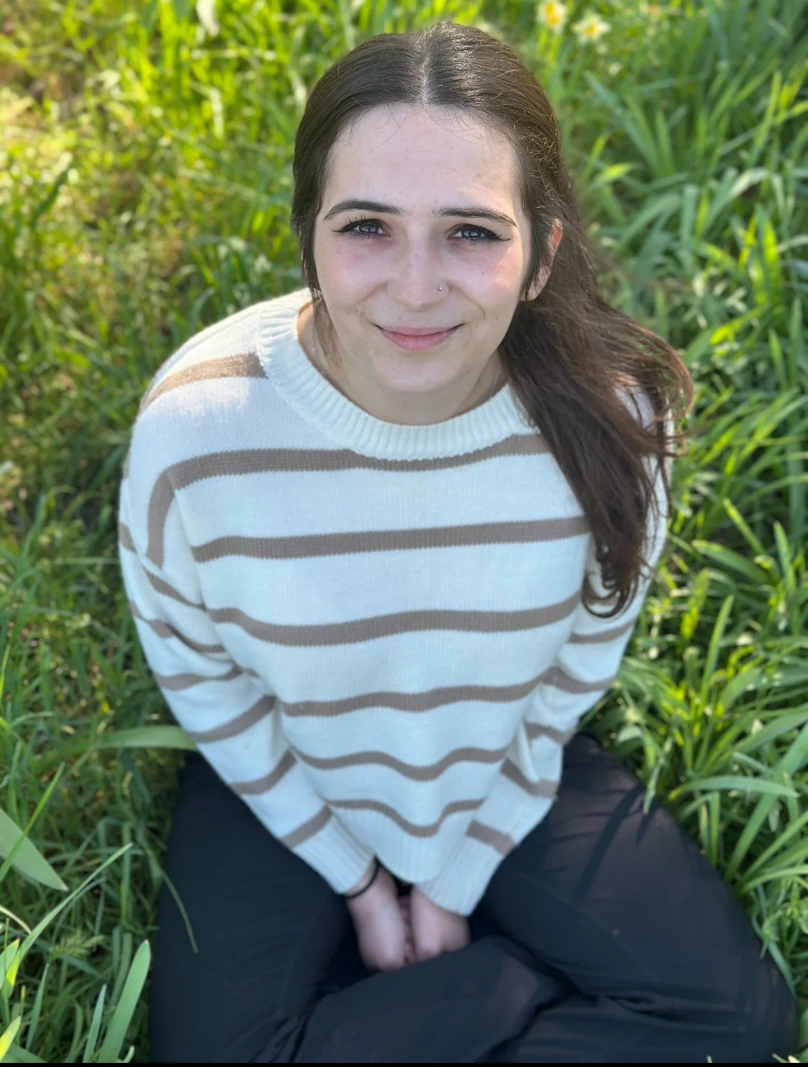 A young woman with dark brown hair, blue eyes, and a nose piercing, sitting outdoors among green plants, wearing a cream and brown striped sweater and black pants, smiling at the camera on a sunny day.