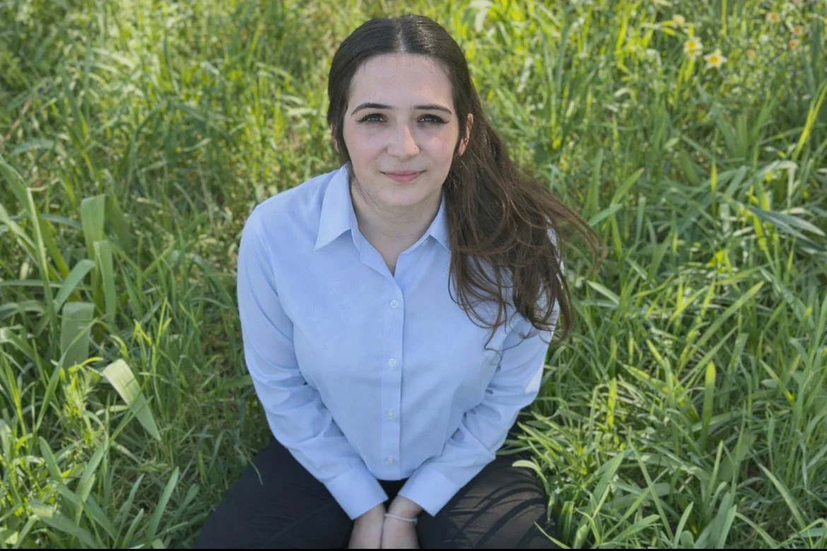 Young woman with long brown hair, wearing a light blue shirt, sitting in a field of tall green grass, looking at the camera.