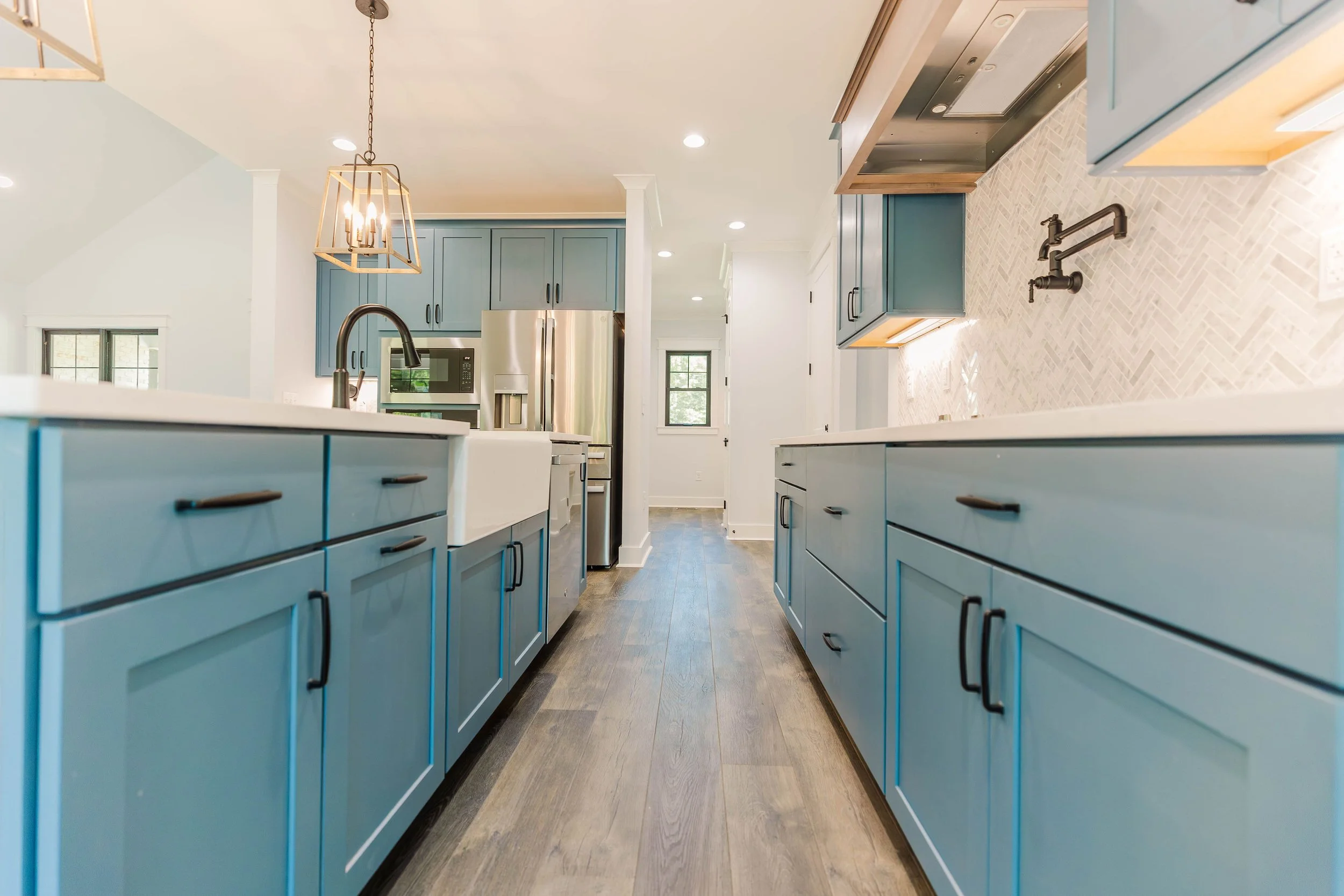 Modern kitchen with blue cabinets, a white countertop, a stainless steel refrigerator, and a herringbone tiled backsplash. There is a black matte faucet and a gold pendant chandelier.
