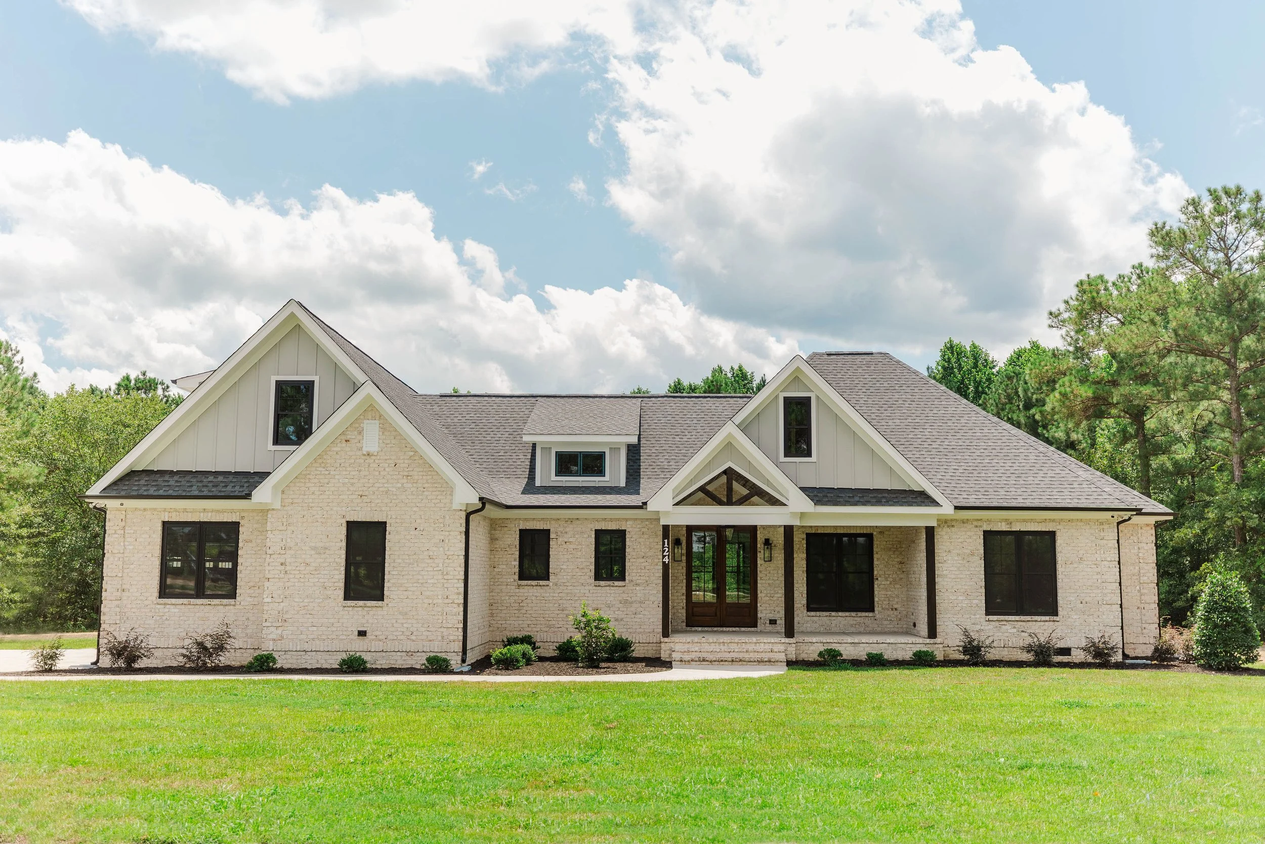 Front view of a modern house with a brick exterior, dark gray roof, and large windows, surrounded by a green lawn and trees, under a partly cloudy sky.