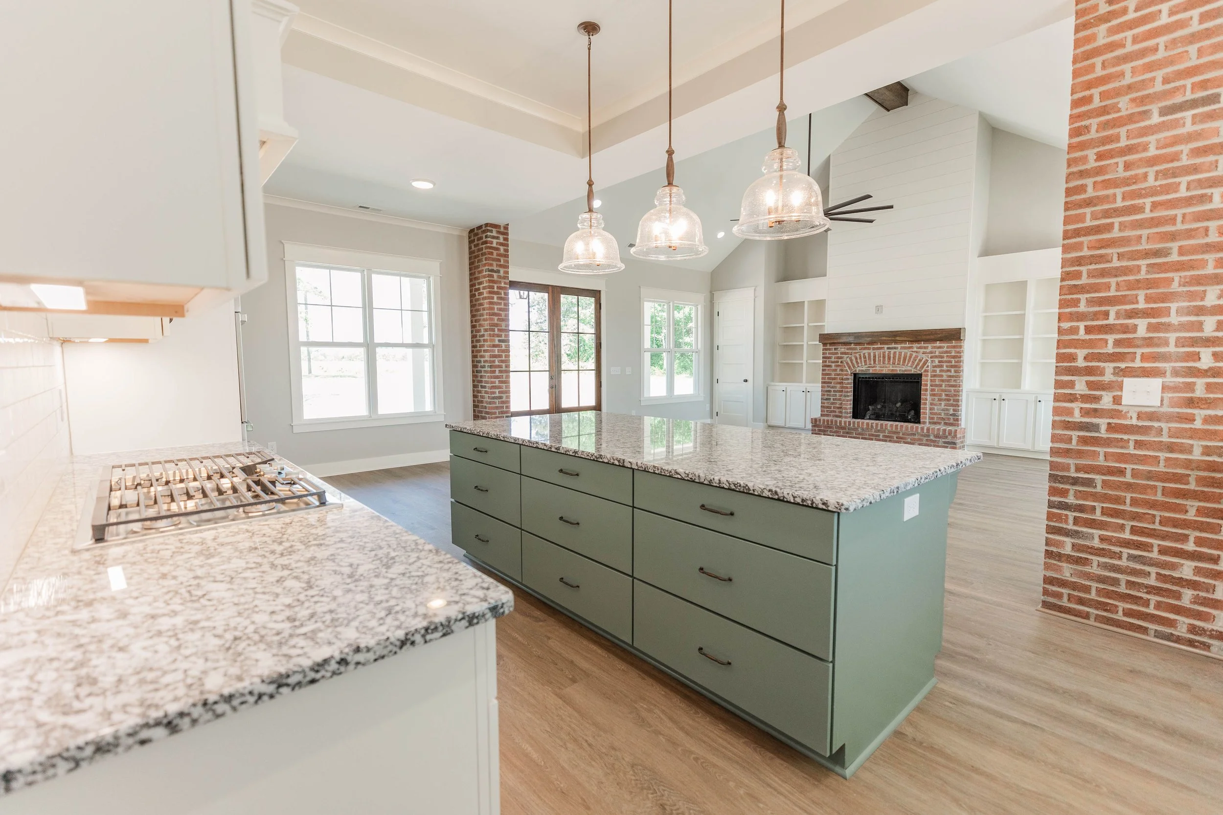 Open-concept kitchen with green island, granite countertops, brick fireplace, white shelves, large windows, and pendant lights.