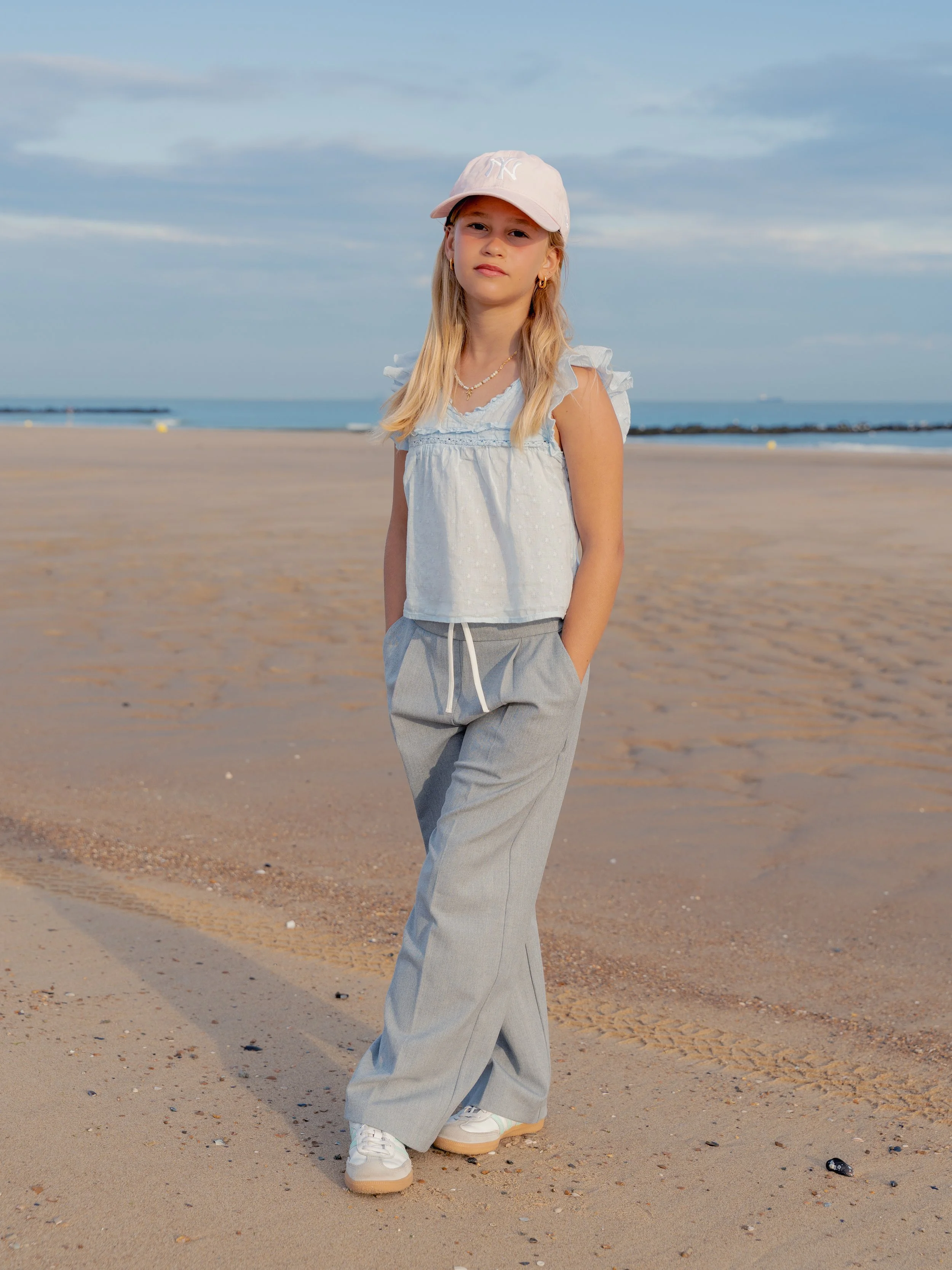 Een jong meisje staat op het strand tijdens zonnig weer. Ze draagt een lichtblouse, grijze wijde broek, witte sneakers, een roze pet en heeft lang blond haar.