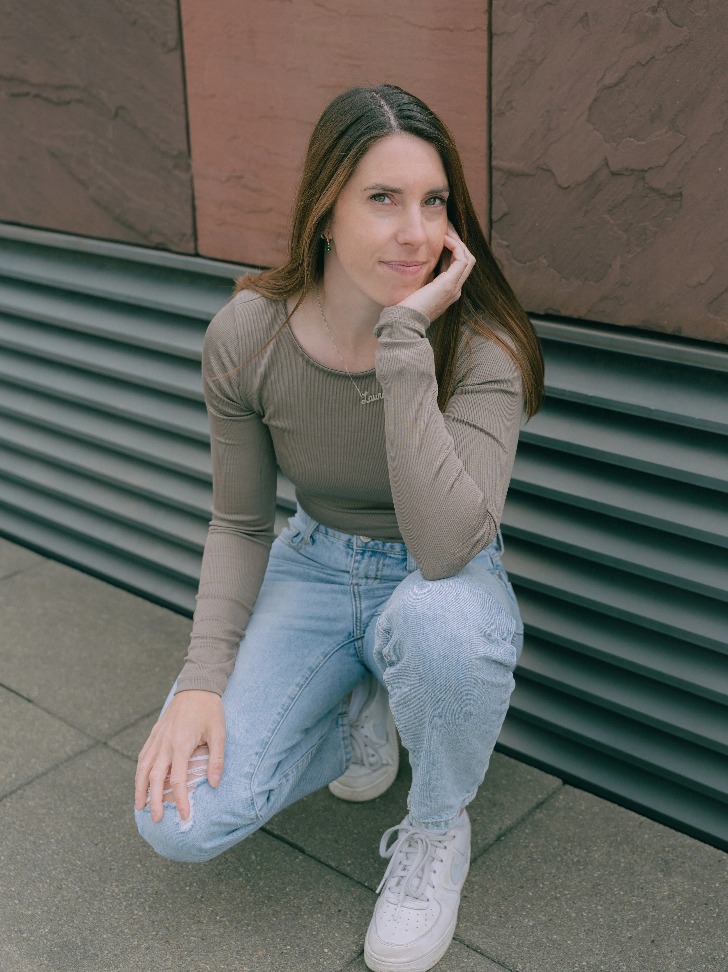 Een vrouw in een beige lange mouwen shirt en lichtblauwe high-waisted jeans met witte sneakers, geknield op straat, met een achtergrond van een muur met roestkleurige en grijze panelen.