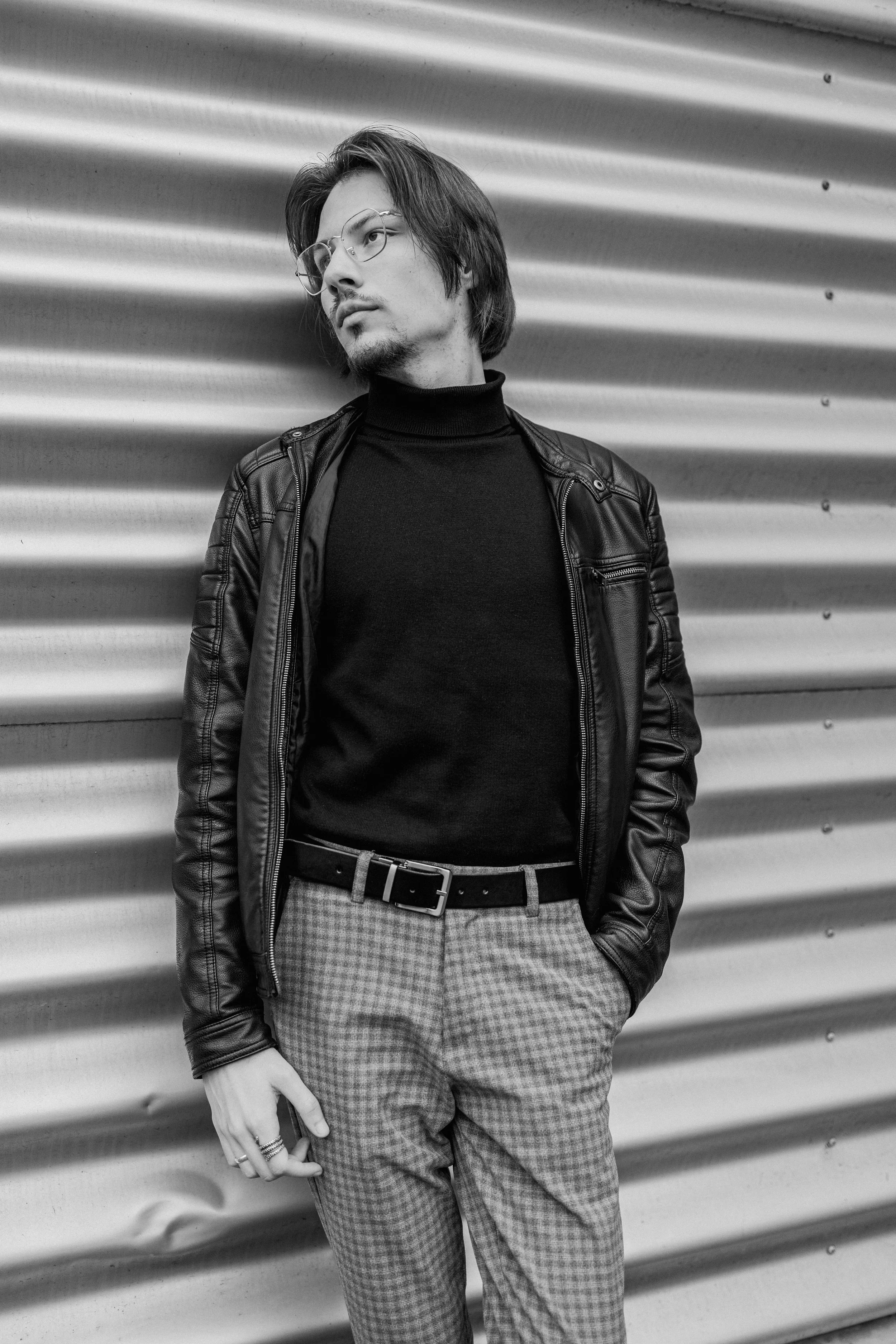  man in zwart leren jasje en geruite broek leunt tegen metalen muur in zwart-wit foto