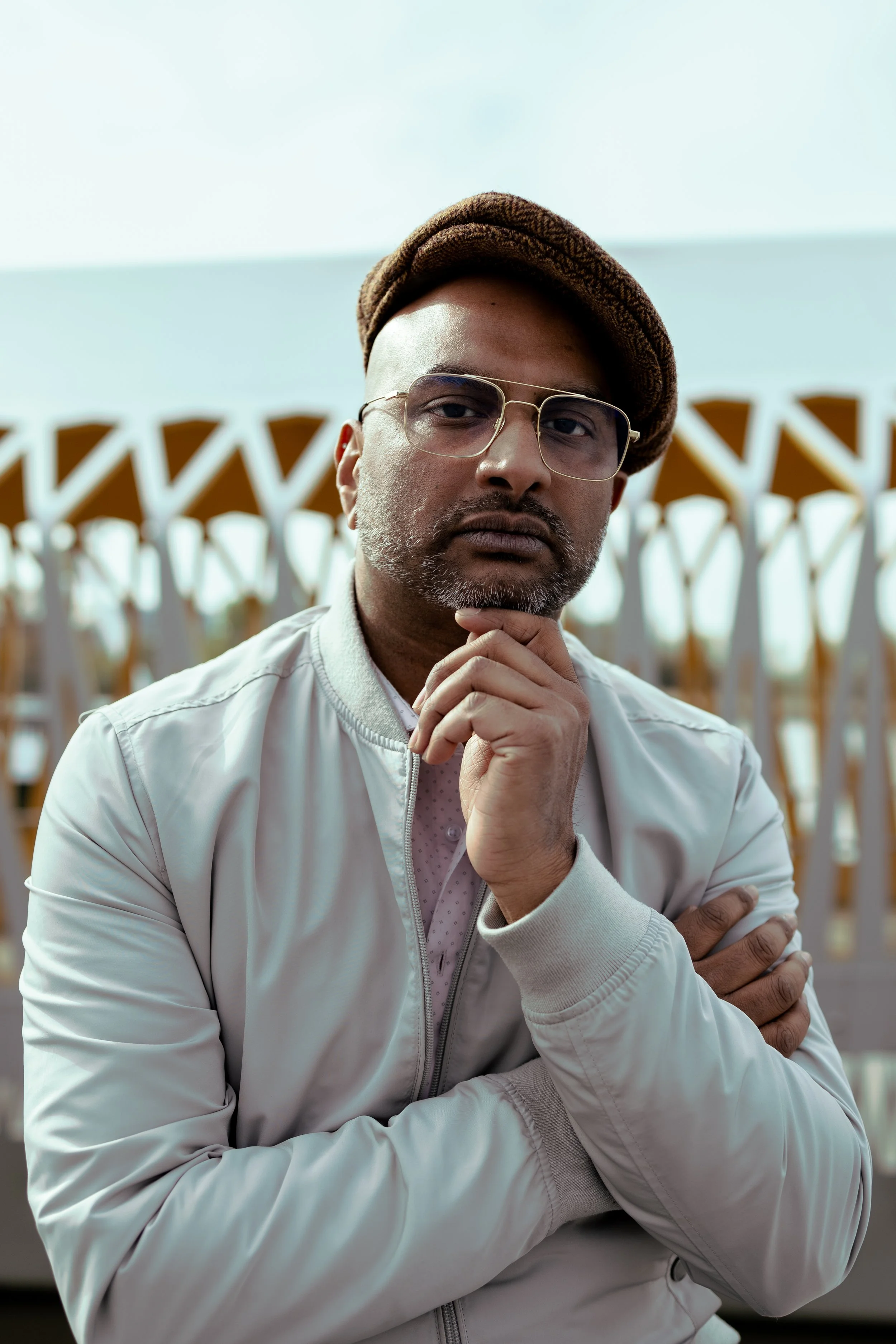 Portret van een man met een bril, een pet en een lichtkleurige jas, met een abstracte achtergrond.