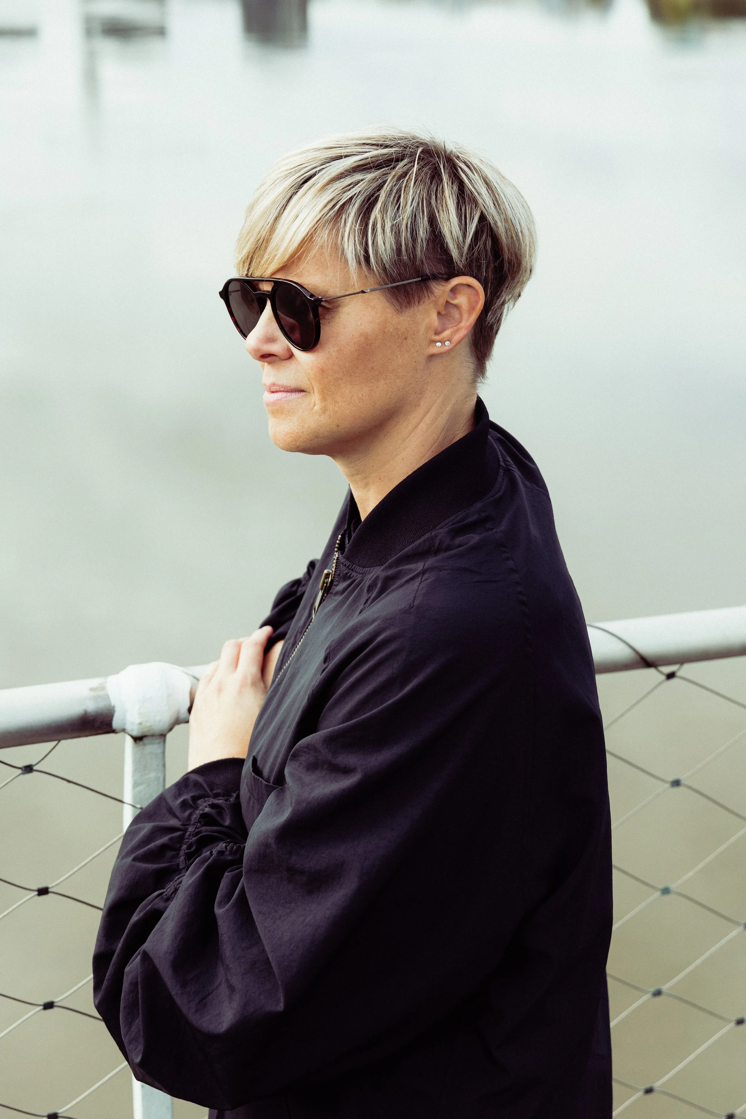 Een vrouw met korte, gespikkelde highlights haar haar en donkere zonnebril staat aan een hek met water op de achtergrond.