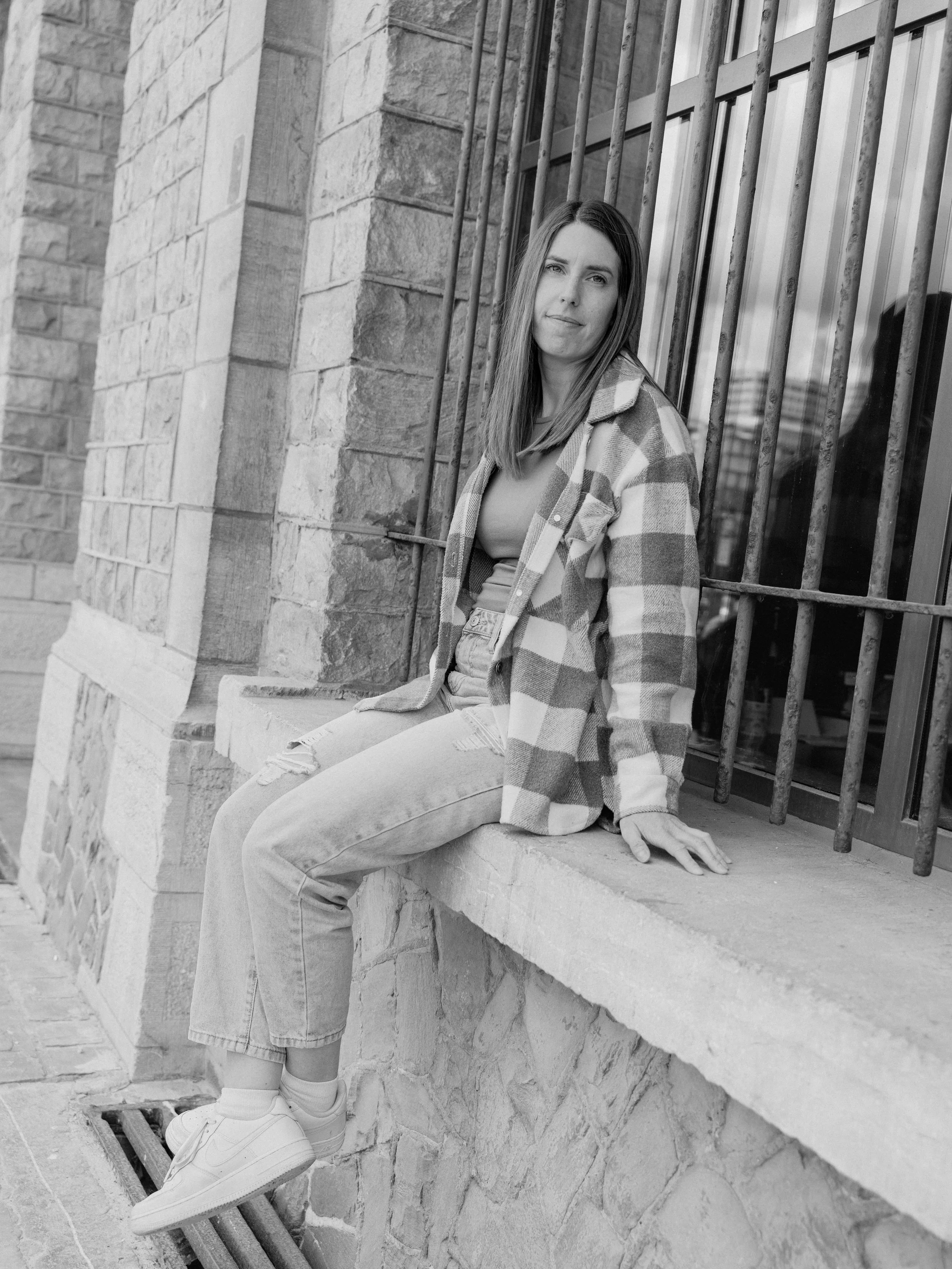 Een jonge vrouw met lang haar, zittend op een stenen rand naast een traliehek, gekleed in een plaid jas, een T-shirt en ripped jeans, in een zwart-wit foto.