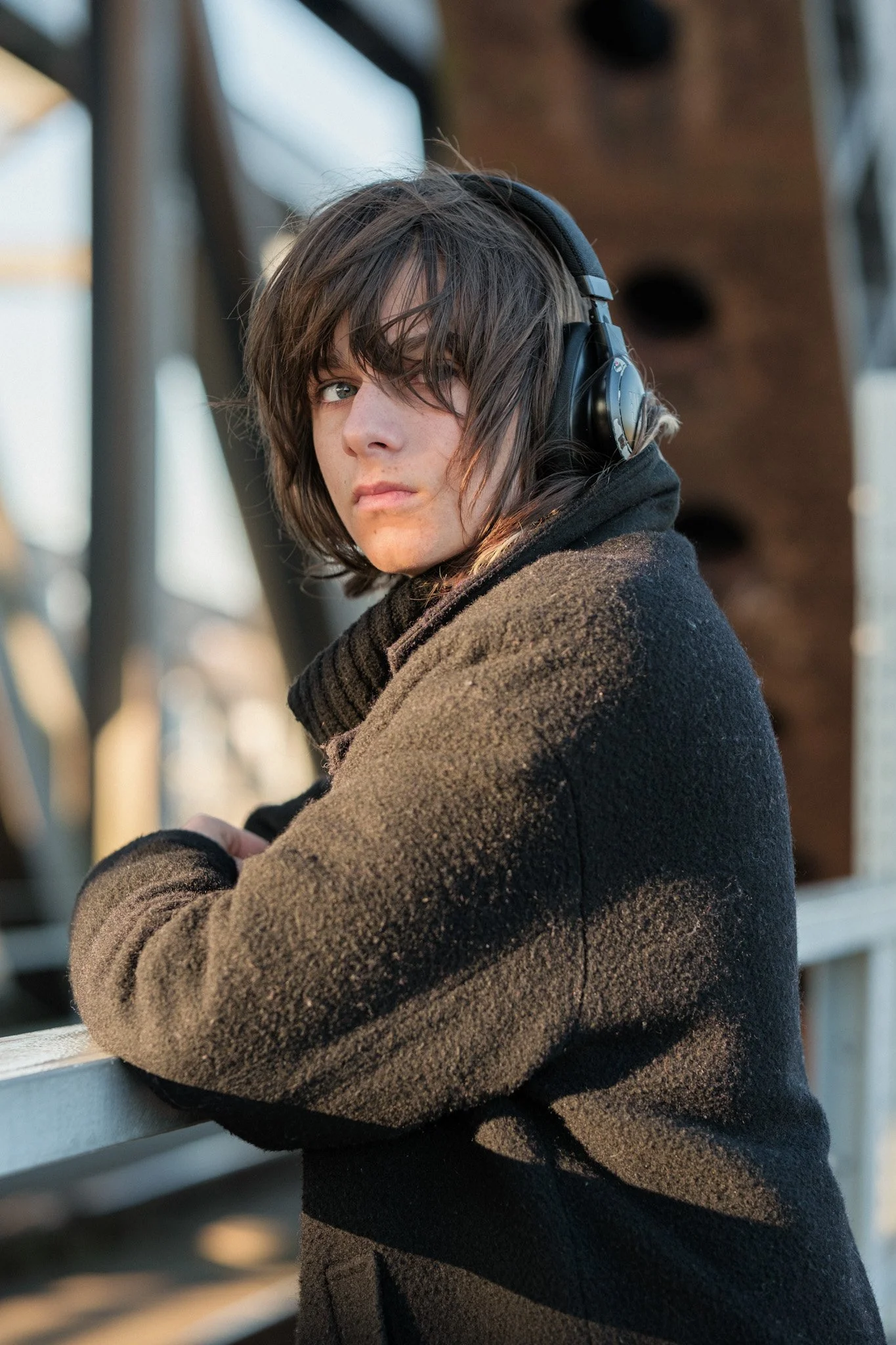 Jonge persoon met donkere haar die naar camera kijkt, draagt zwarte koptelefoon en een donkere jas, leunt op een witte reling, met industriële structuur op achtergrond.