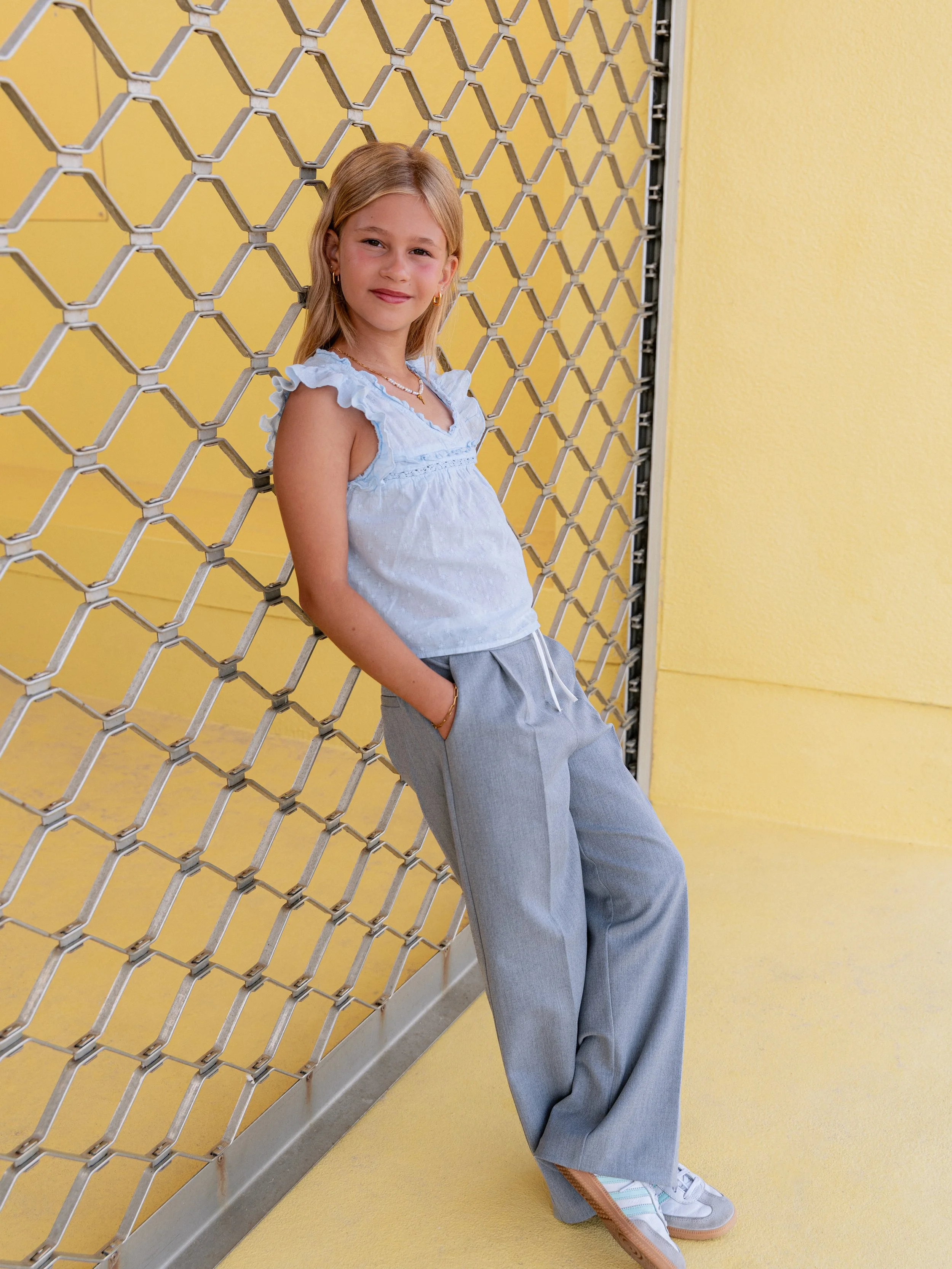 Een jonge meid leunt tegen een metalen hekwerk, gekleed in een lichtblauwe top, grijze wijde broek en witte sneakers, staand tegen een gele muur.