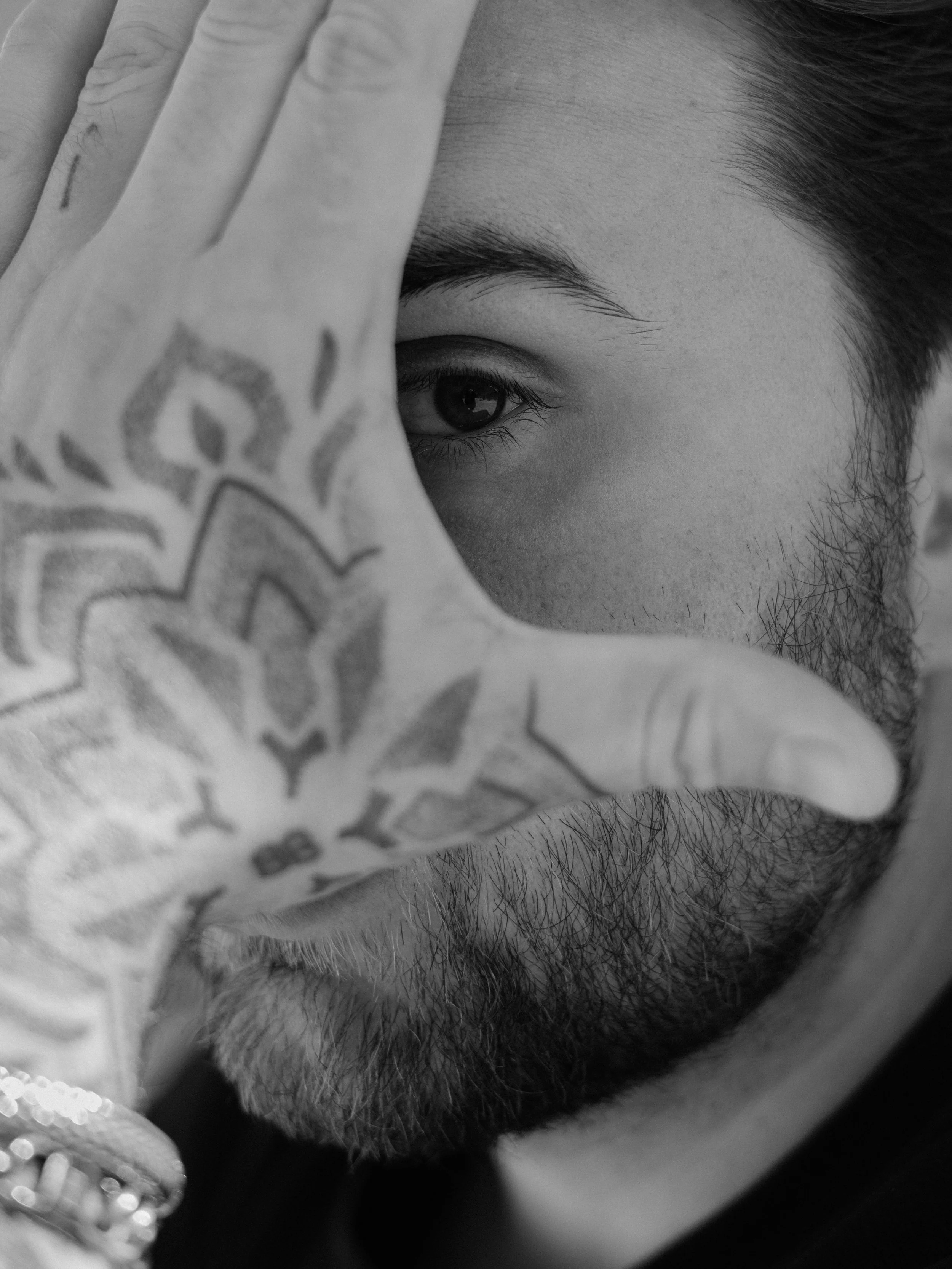 Close-up van een man met een gezichtshaar, die door zijn hand met een tattoeage het oog en een deel van het gezicht zichtbaar maakt.