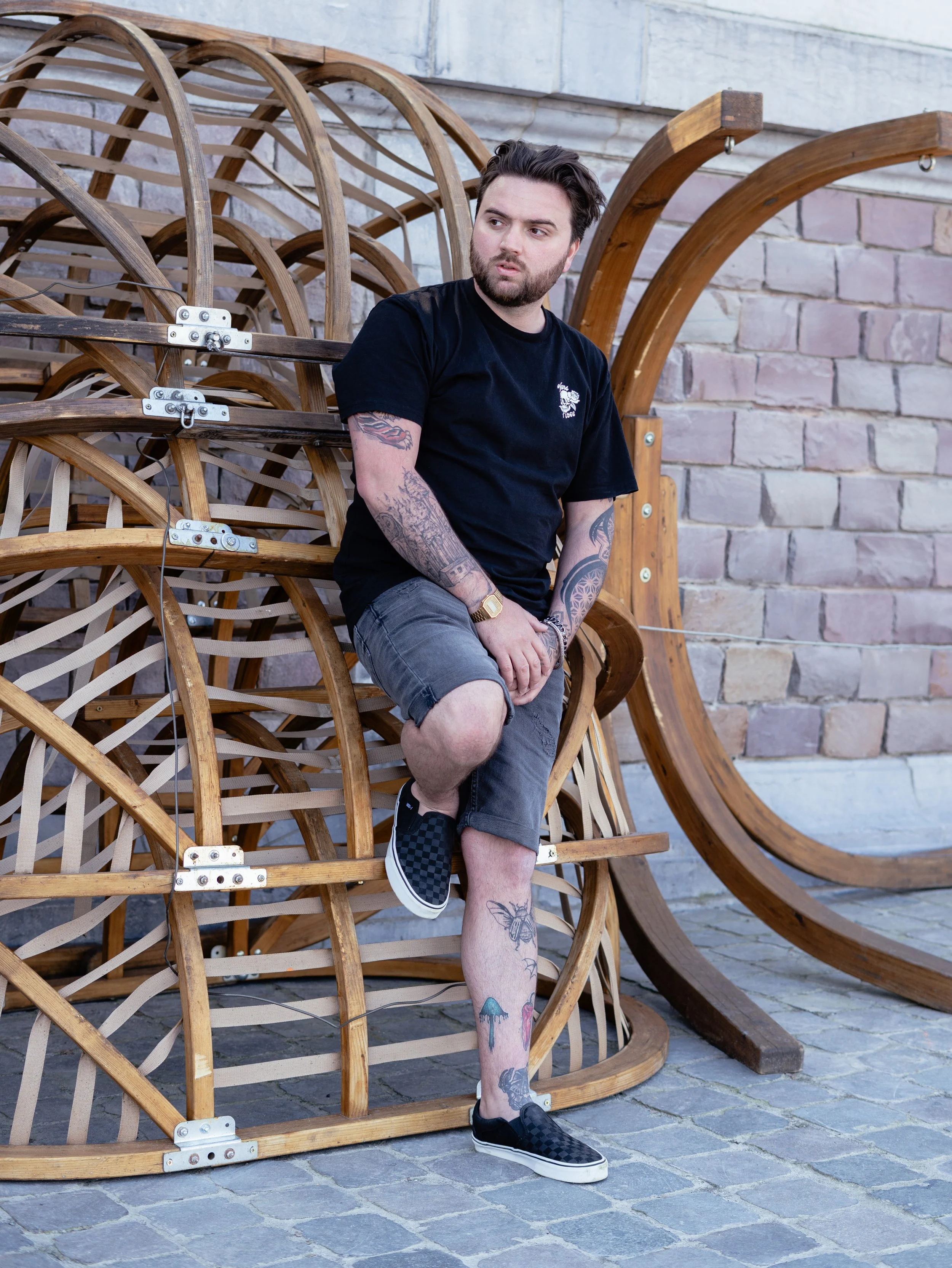 Man met tattoo's zittend op houten kunstwerk, gelegen tegen bakstenen muur, draag zwart T-shirt en shorts, met sneakers.