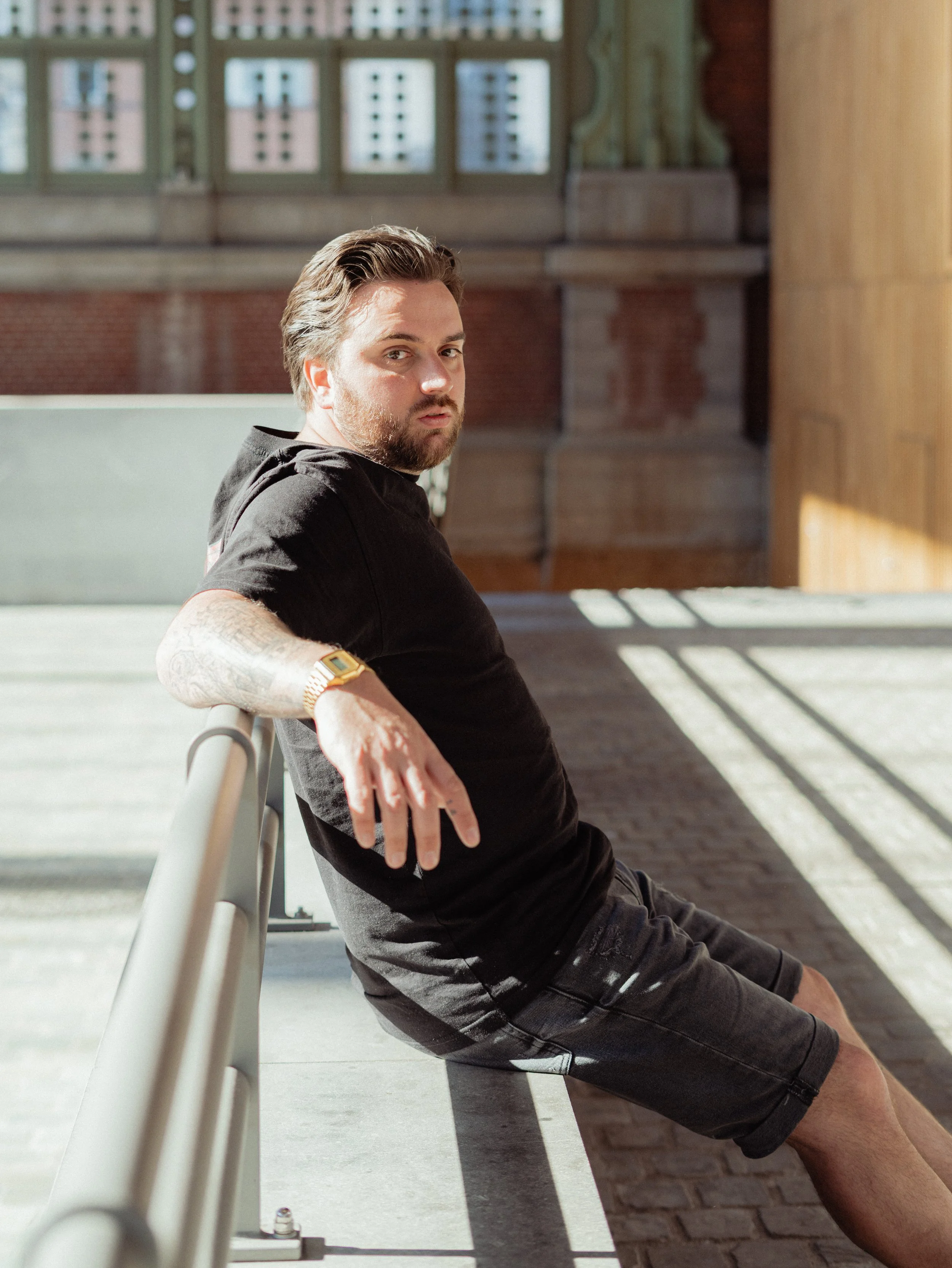 Mannelijke persoon met tattoo op arm, zittend op een bank in een gebouw met veel ramen, gekleed in een zwart T-shirt en zwarte broek, met natuurlijke verlichting en schaduwen op de vloer.