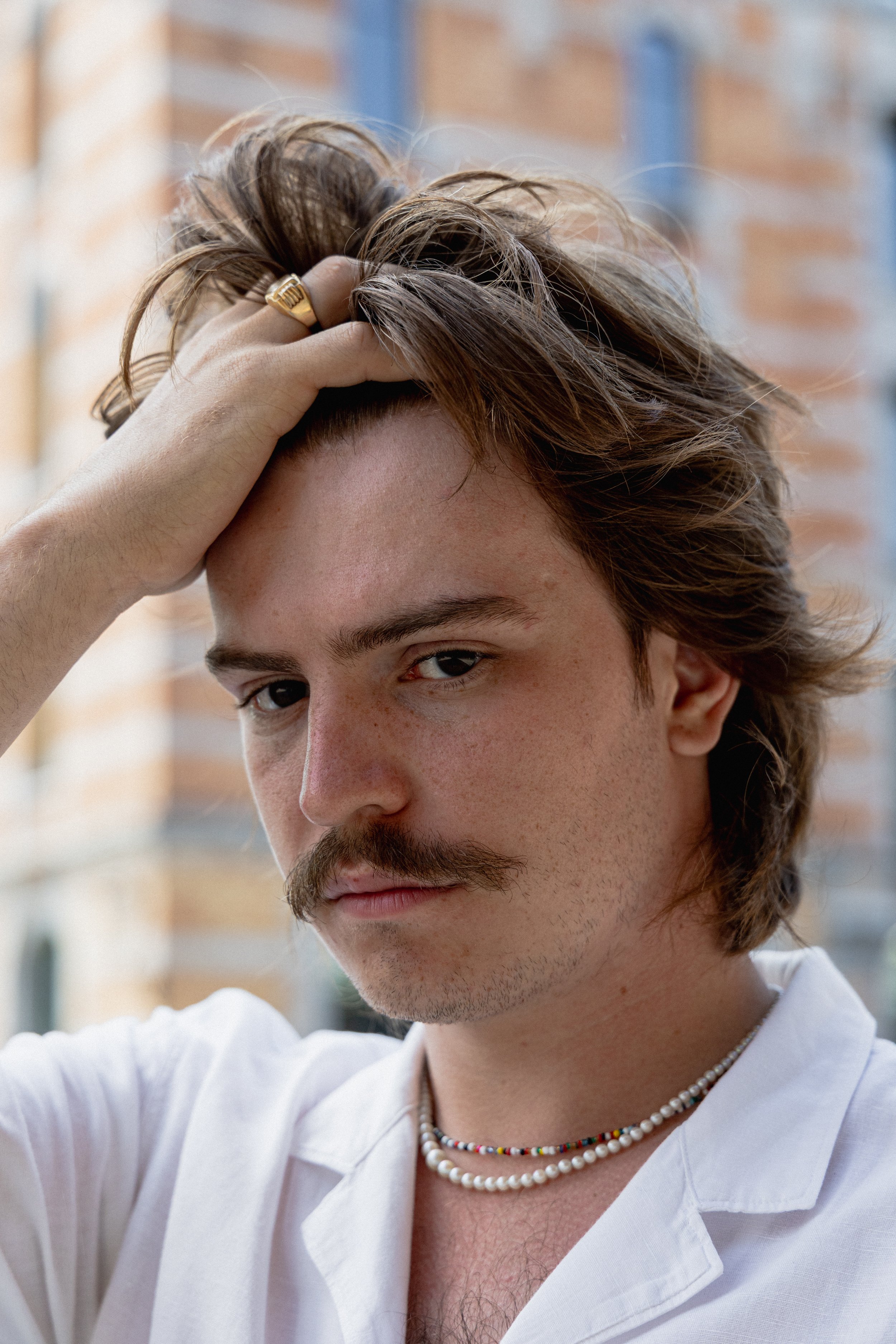 Portret van een jonge man met een lichte huid, een snor en een lichte baard, die rust op zijn hoofd met zijn hand. Hij draagt een witte blouse, een parelketting en een gekleurde ketting. De achtergrond toont een gebouw met bakstenen muren.