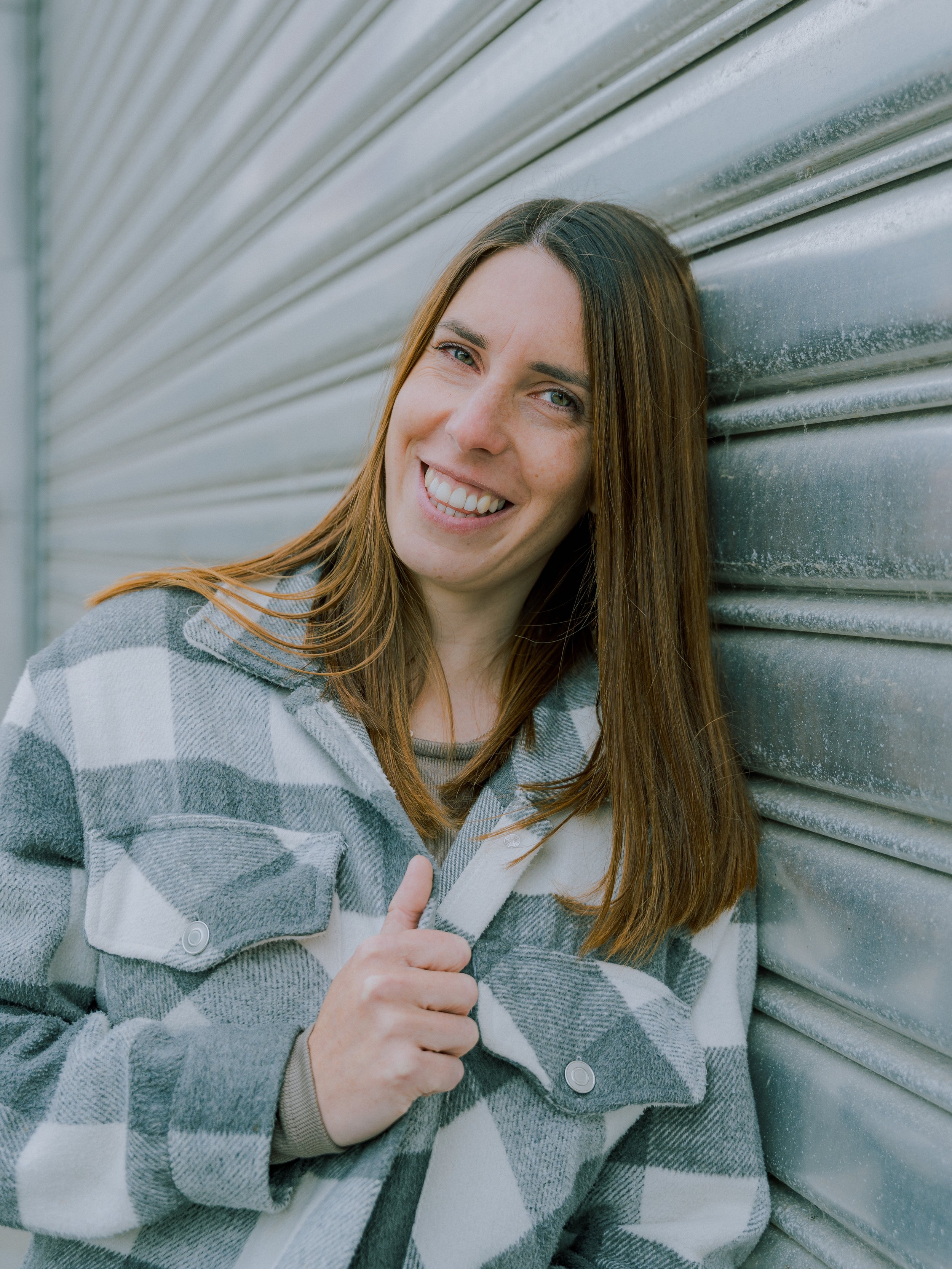 Een lachende vrouw met bruin haar in een grijs-wit geruit jasje die naast een metalen loods staat.