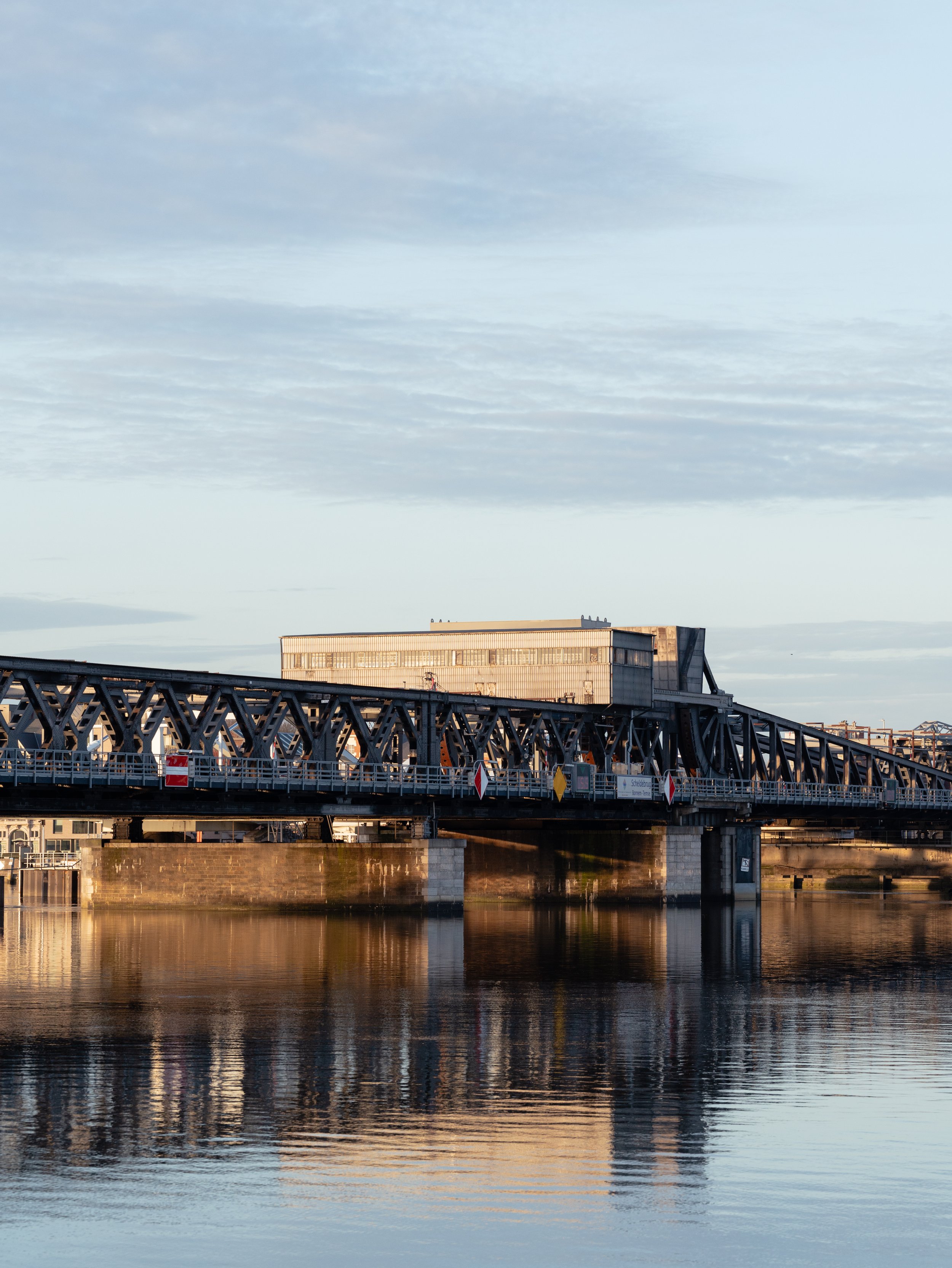 Foto van een brug over een rivier met een modern gebouw op de achtergrond tijdens zonsondergang.