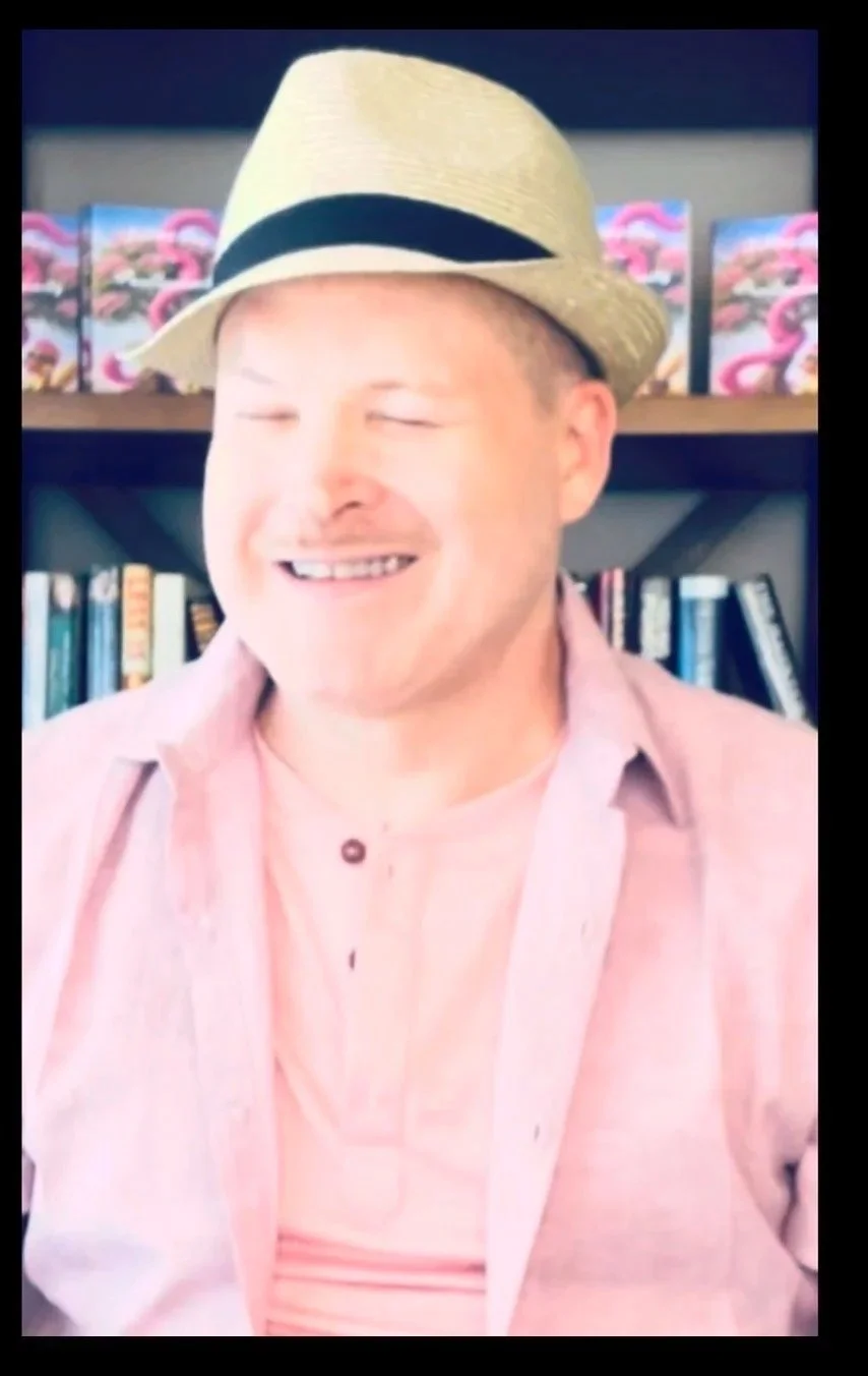 A man smiling with eyes closed, wearing a beige fedora with a black band and a pink shirt, standing in front of a bookshelf with books and decorative flamingo boxes.