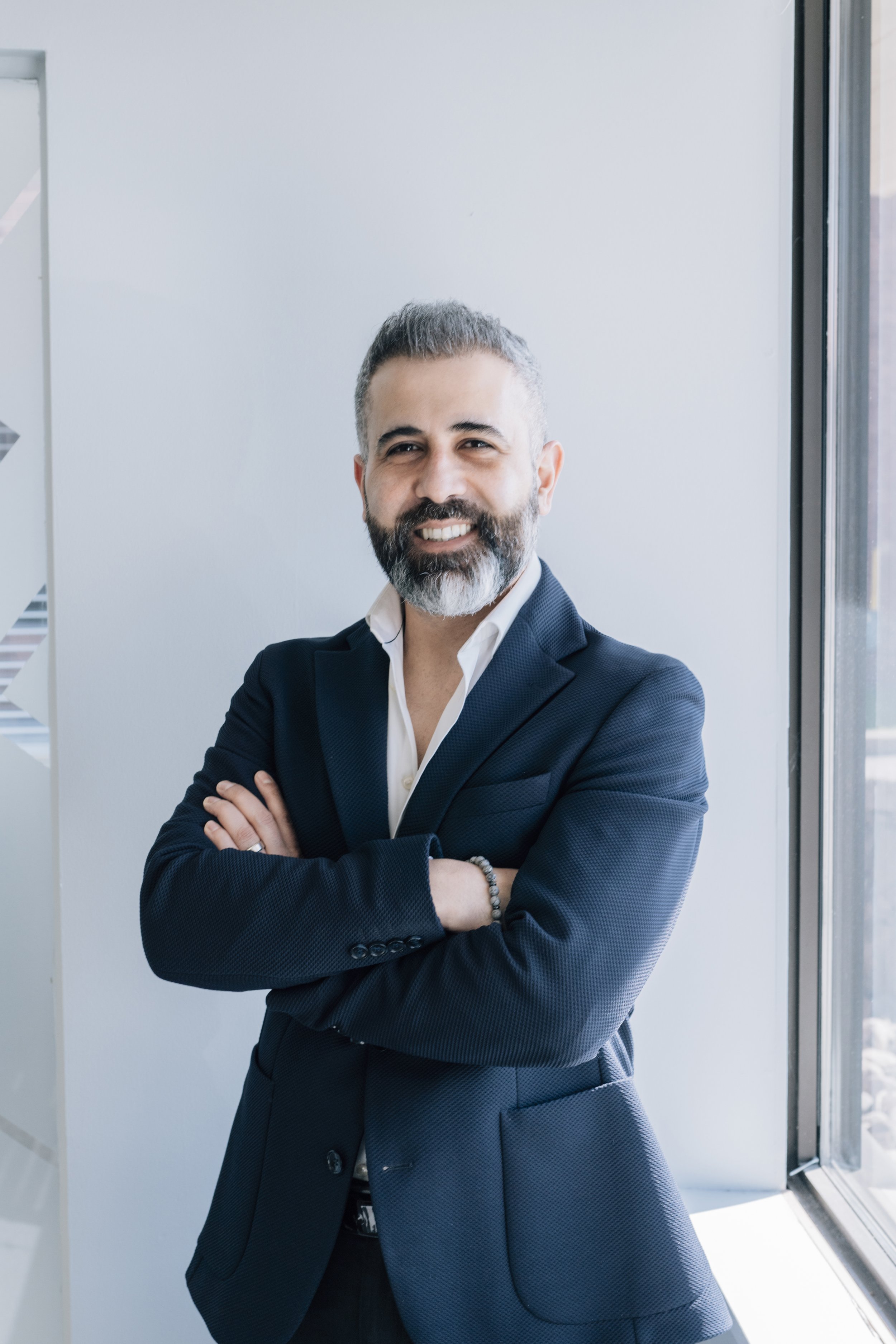 A confident man in a dark blazer standing with arms crossed, smiling, near a window in a modern office.