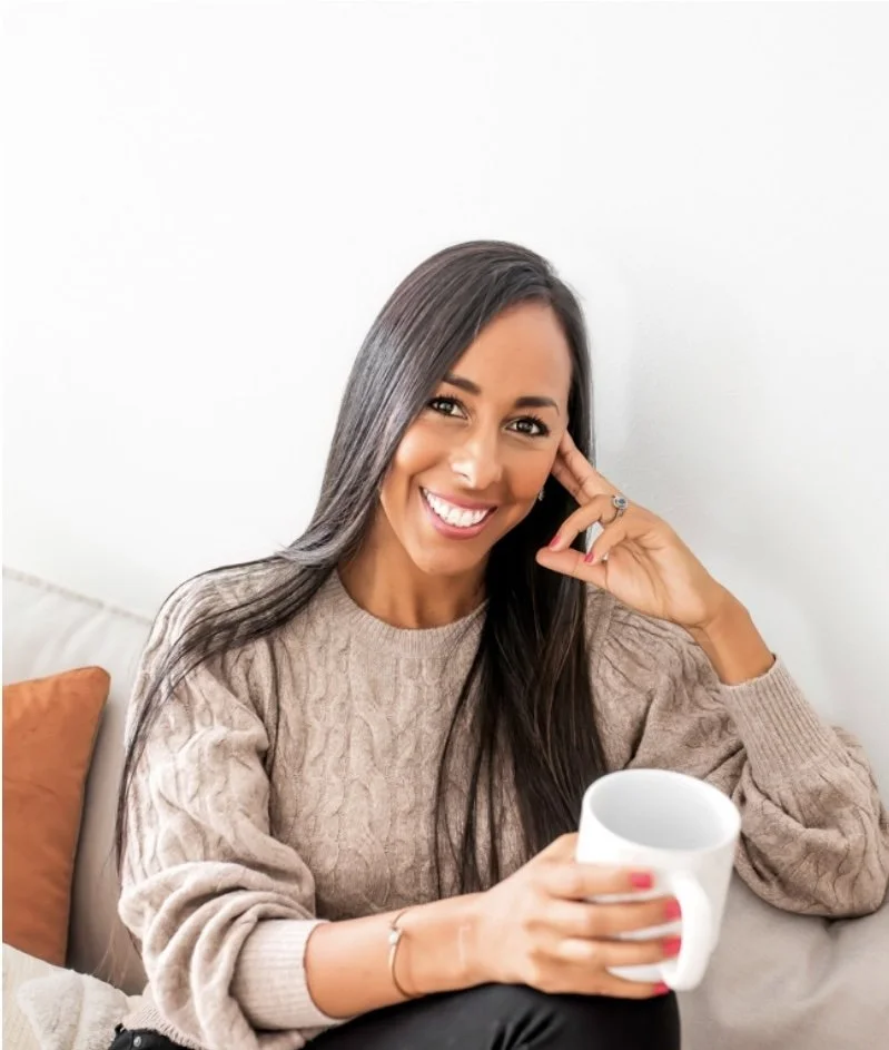 A woman with long dark hair smiling and holding a white mug, sitting on a beige couch with a brown pillow beside her, in a cozy setting.