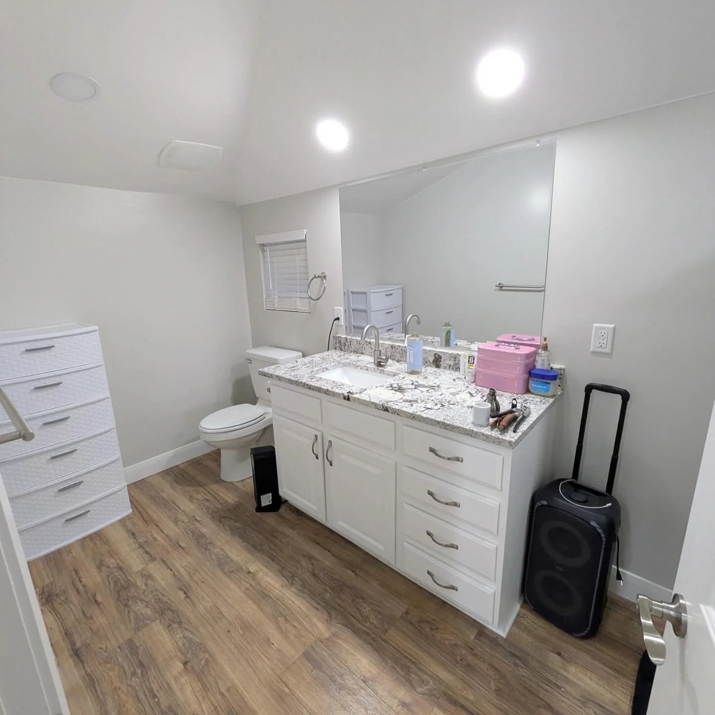 A bathroom with white walls, a granite countertop with two sinks, a large mirror, and wooden flooring. There is a white cabinet, a toilet, and a dresser with multiple drawers. Items on the counter include pink boxes, bottles, and toiletries. A black speaker on the floor, a small white chest of drawers, and a window with blinds are also visible.