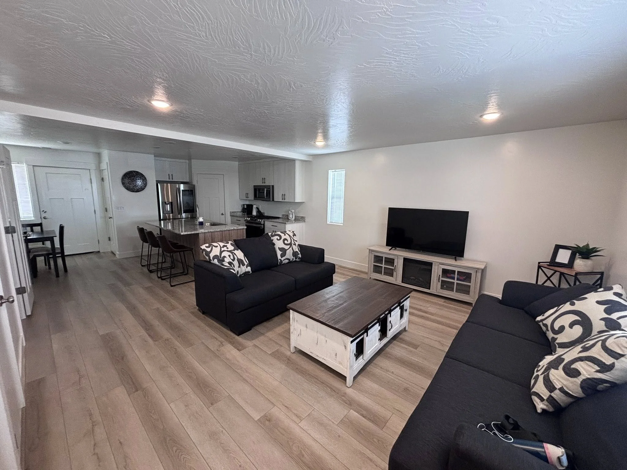 Living room with two black sofas with patterned pillows, a wooden coffee table, a TV on a white TV stand, and an open kitchen with a breakfast bar and barstools.