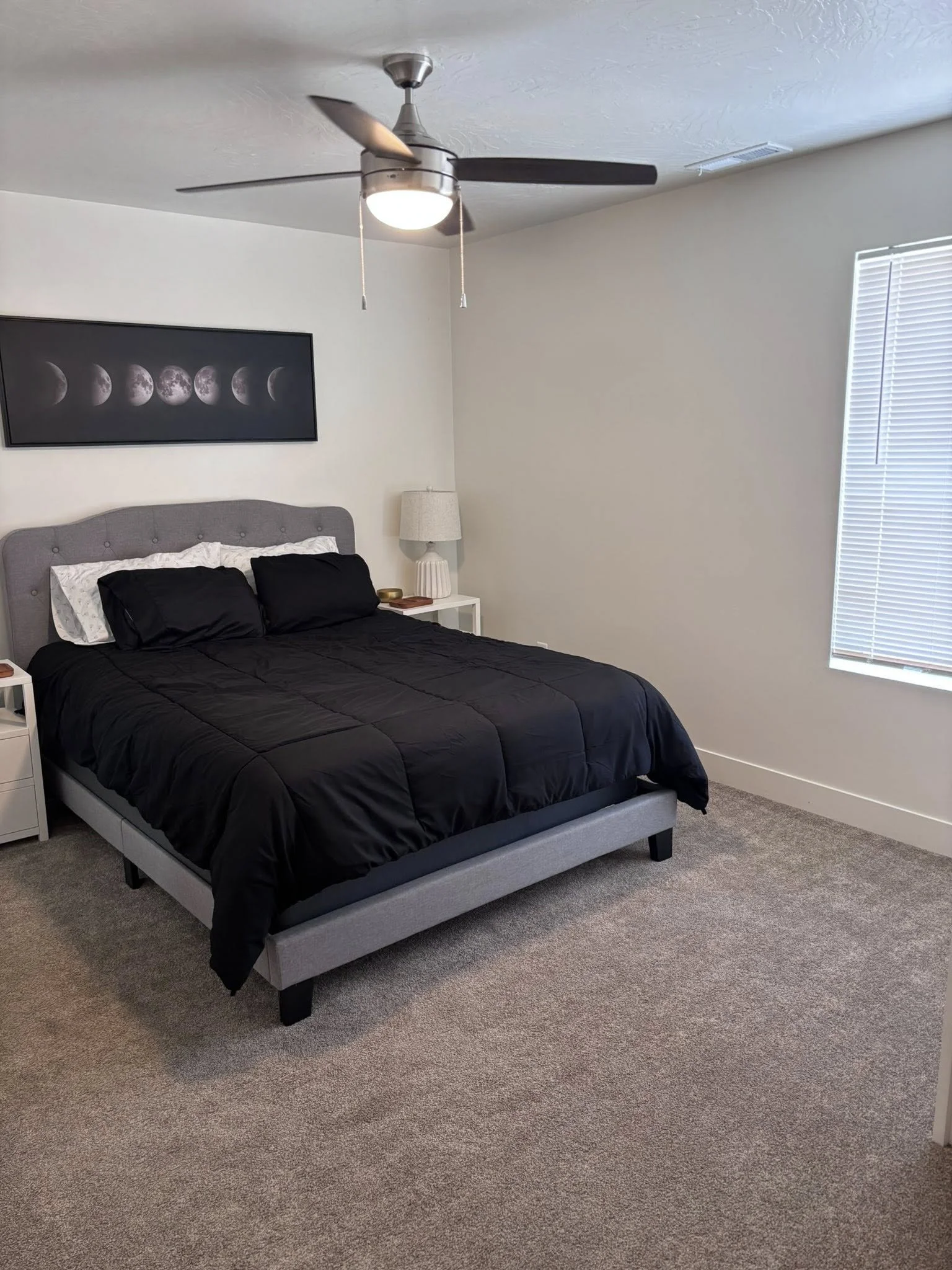 A bedroom with a gray bed frame, black bedding, and two pillows. There are white nightstands with lamps on each side of the bed, and a framed moon phase poster on the wall behind. A ceiling fan with a light is above, and a window with blinds on the right.