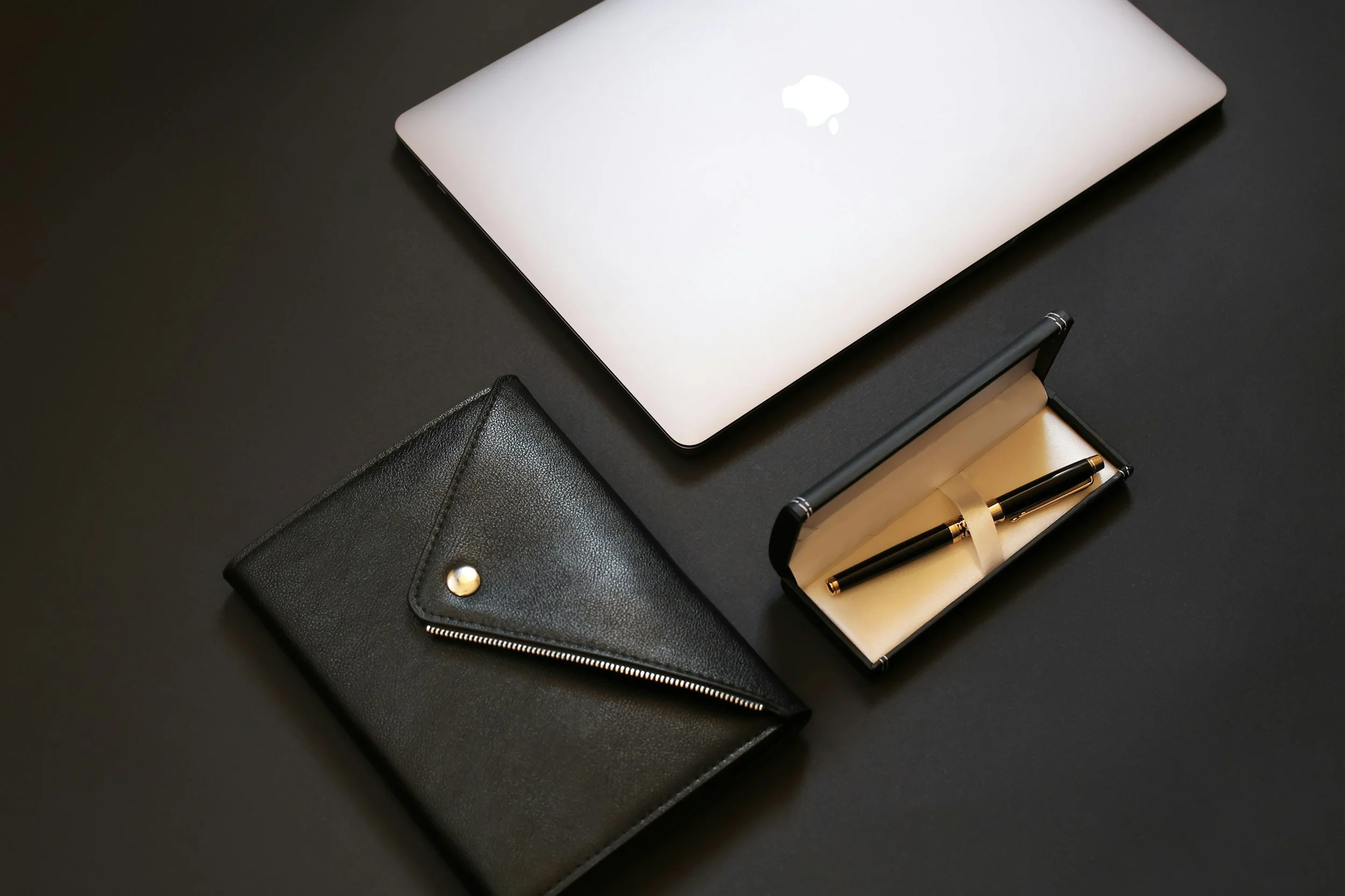 A closed silver MacBook laptop on a black surface, with a black leather wallet and a black and gold pen in a black and gold gift box nearby.