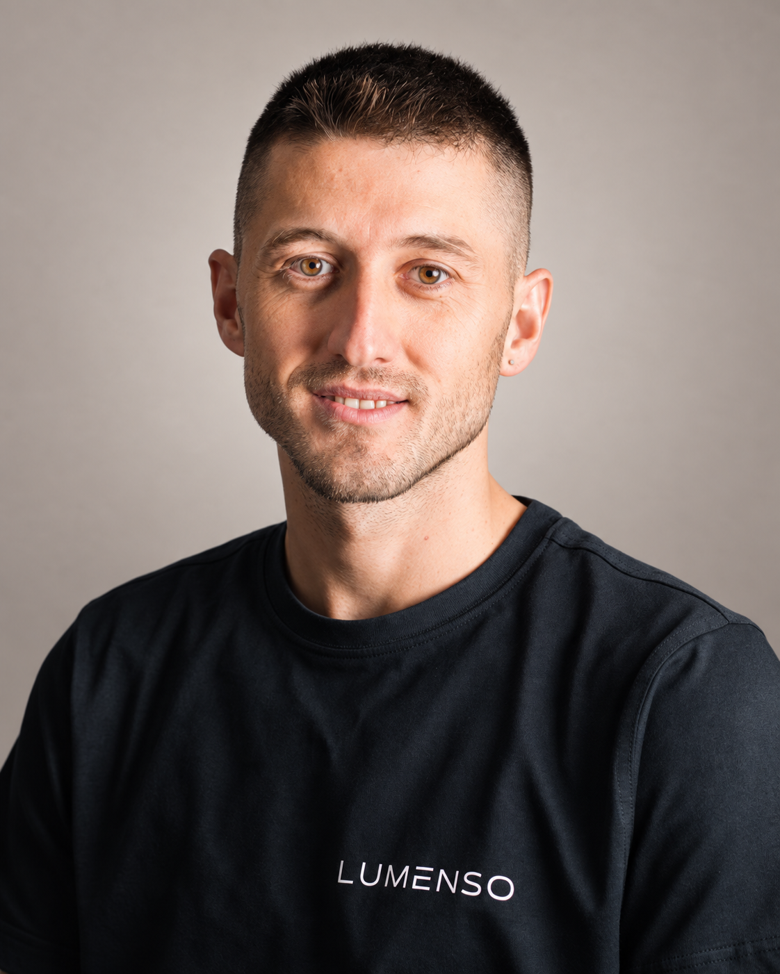 Ein junger Mann mit kurz geschnittenem Haar trägt ein schwarzes T-Shirt mit dem Logo »LUMENSO". Er lächelt und schaut in die Kamera, vor einem weißen Hintergrund.