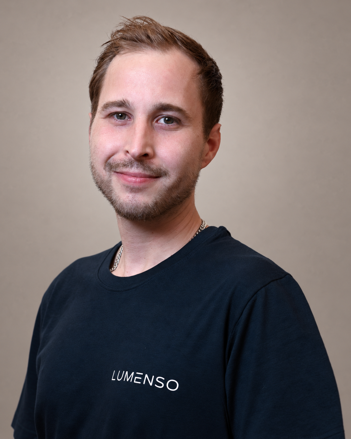 Ein junger Mann mit kurz geschnittenem Haar trägt ein schwarzes T-Shirt mit dem Logo »LUMENSO". Er lächelt und schaut in die Kamera, vor einem weißen Hintergrund.