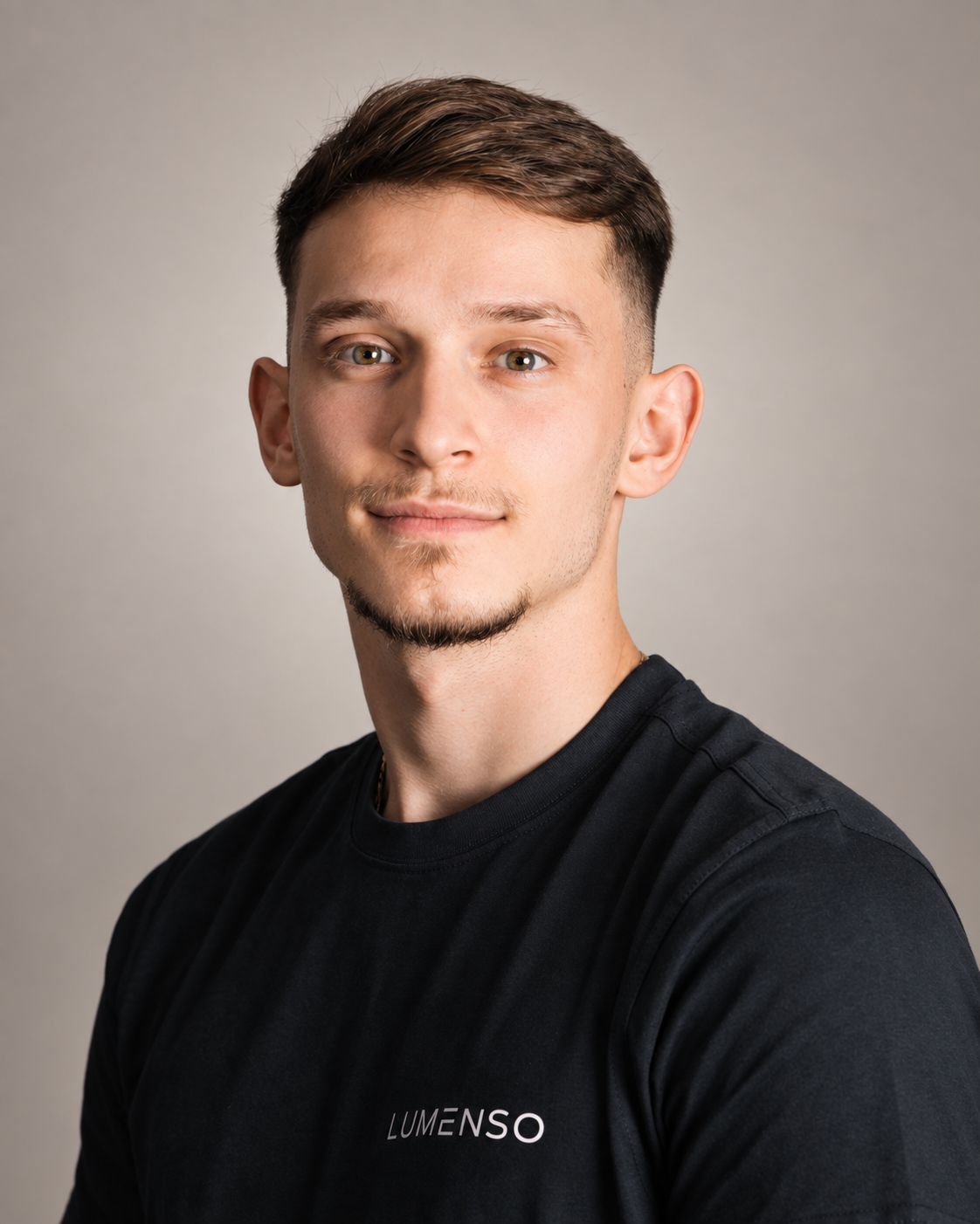 Junger Mann mit kurzem braunem Haar und leichtem Schnurrbart, trägt schwarzes T-Shirt mit Logo 'LUMENSO', blickt lächelnd in die Kamera vor hellem Hintergrund.