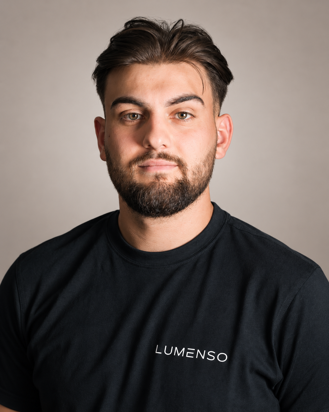Ein junger Mann mit dunklem Haar und Bart trägt ein schwarzes T-Shirt mit dem Schriftzug "LUMENSO" auf der linken Brust. Er blickt direkt in die Kamera vor einem weißen Hintergrund.