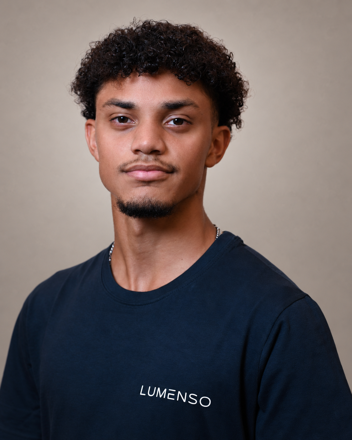 Ein junger Mann mit dunklem Haar und Bart trägt ein schwarzes T-Shirt mit dem Schriftzug "LUMENSO" auf der linken Brust. Er blickt direkt in die Kamera vor einem weißen Hintergrund.