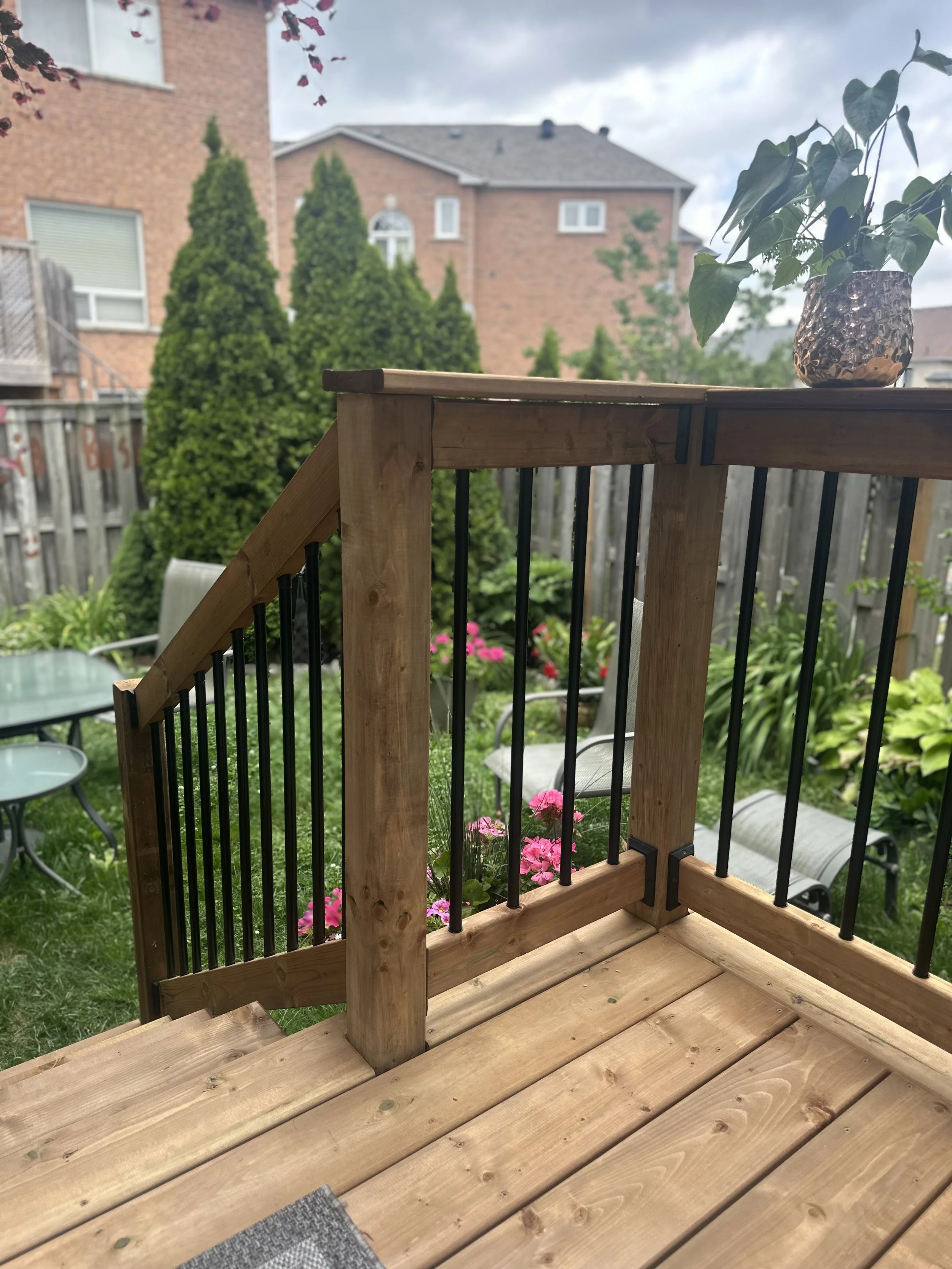 View of a backyard deck with a railing, potted plant, and garden with pink flowers, trees, and neighboring brick houses under cloudy sky.