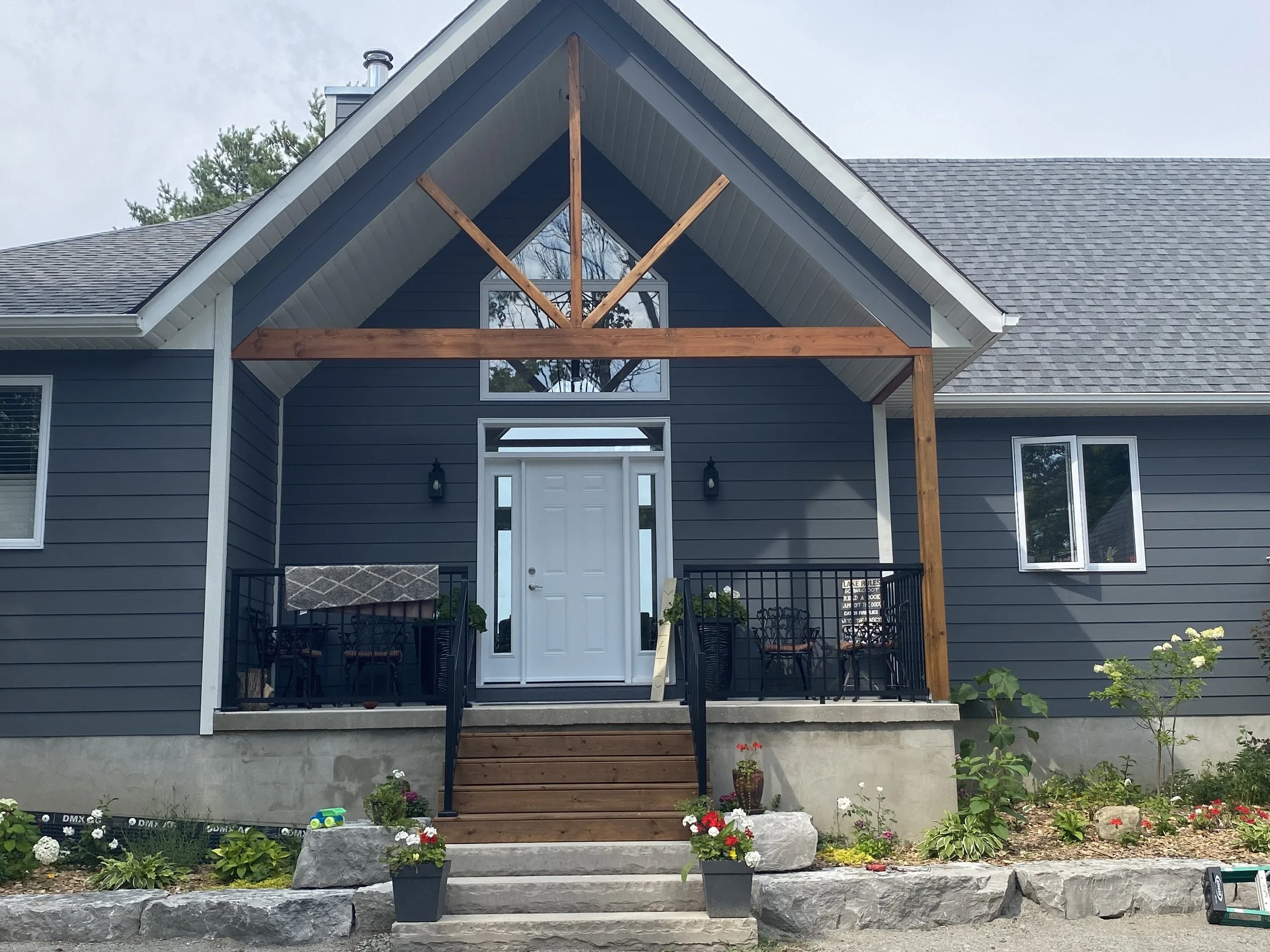 Front of a modern house with blue siding and white trim, a front door, and a porch with wooden beams, railing, and outdoor furniture, landscaped with flowers and plants.