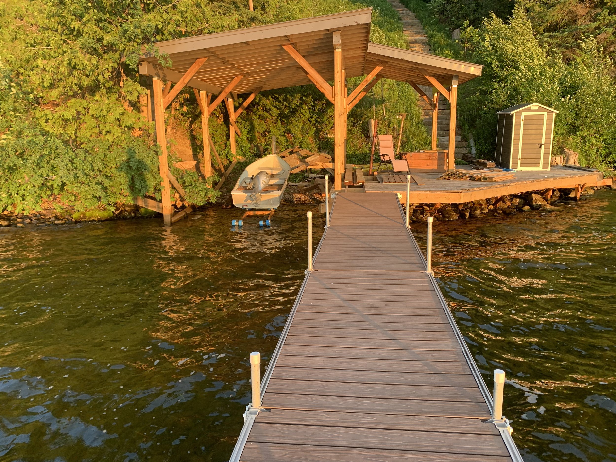 Wooden dock leading to a shoreline cabin with boat shed, boat, and two lounge chairs, surrounded by green trees, during sunset.