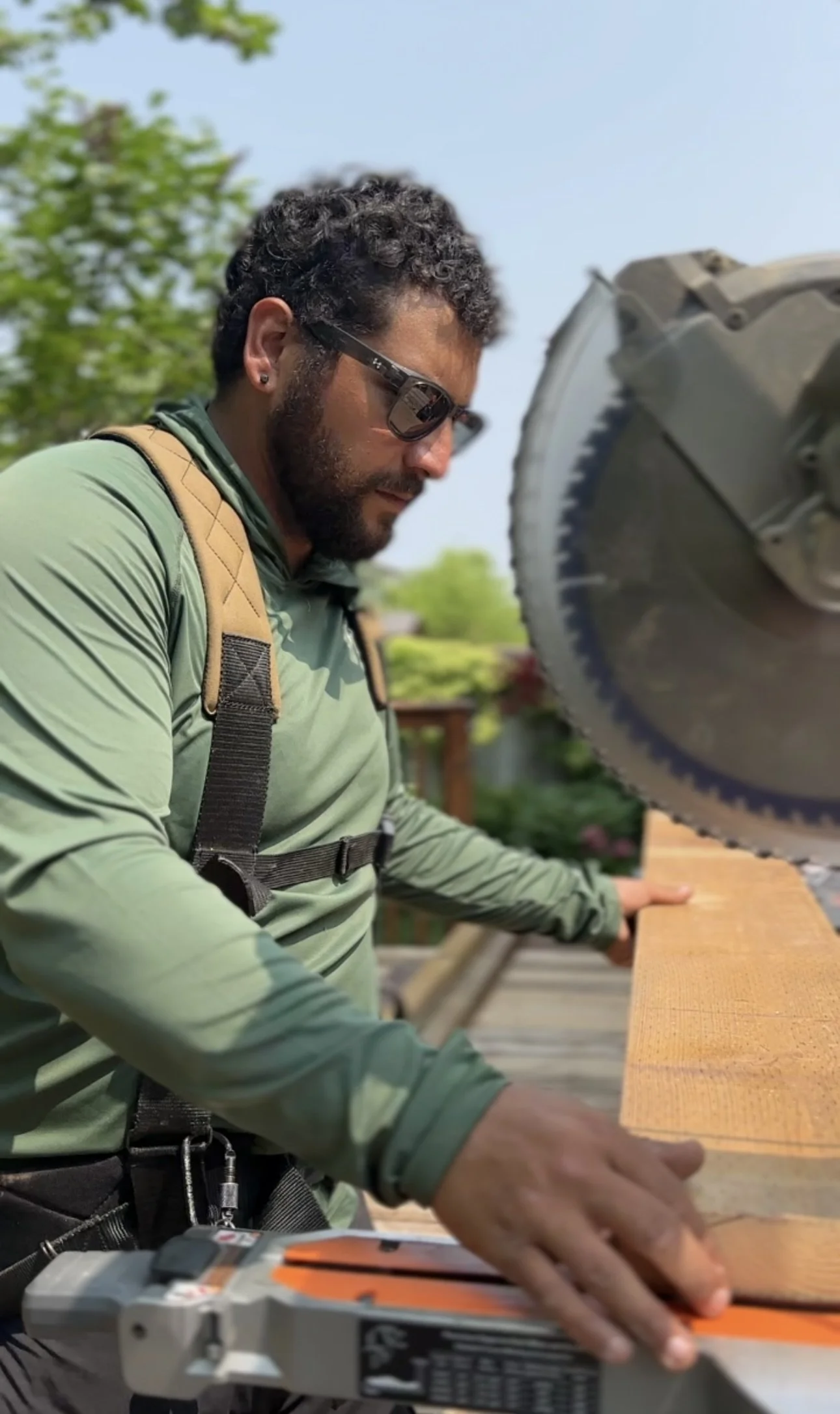 A man with curly hair and sunglasses is working with a compound miter saw in a woodworking project outside. He is wearing a green long-sleeve shirt and a tan backpack, and is holding a piece of wood against the saw's table.