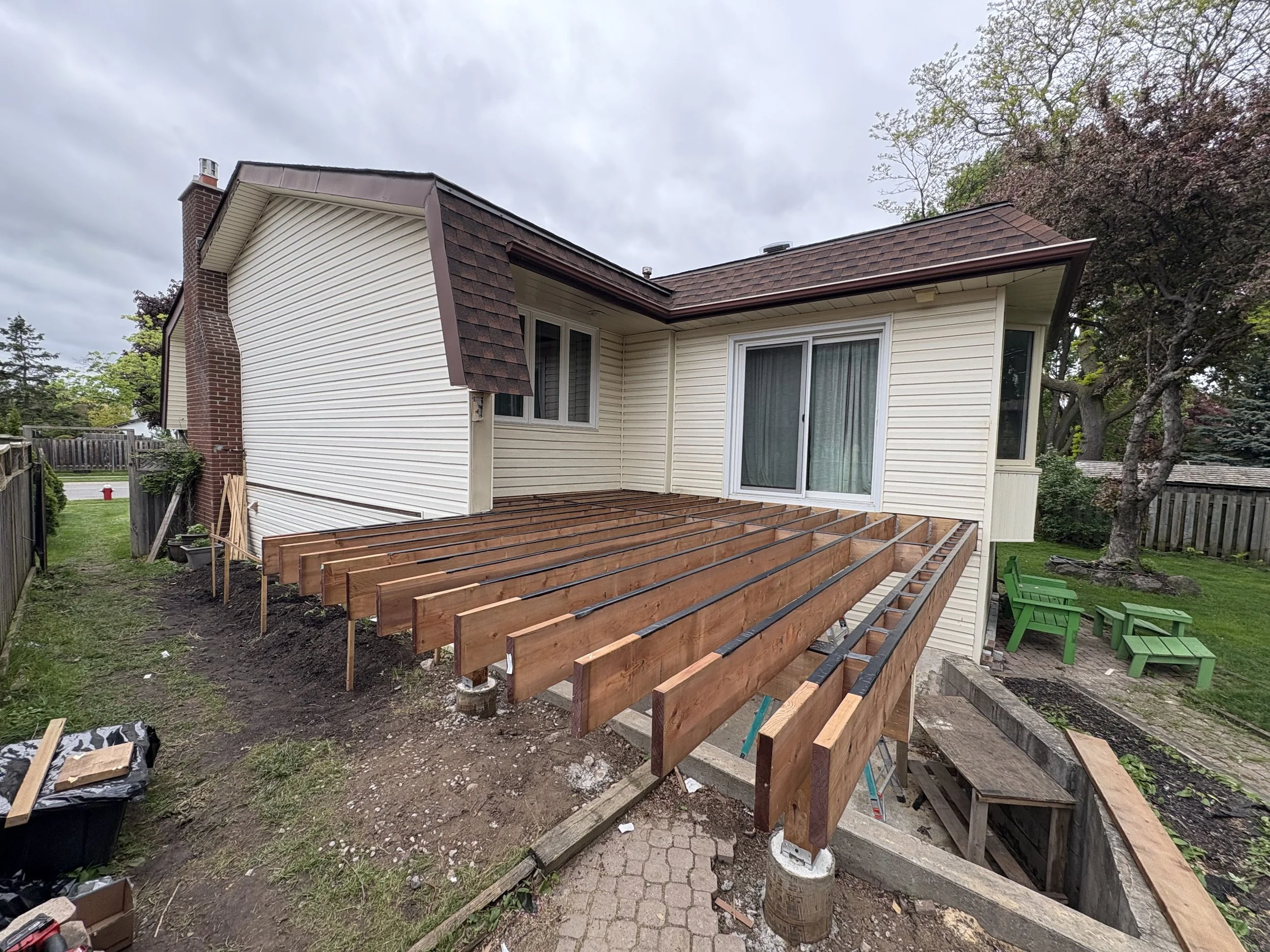 Partially constructed wooden deck attached to the back of a house with beige siding and sliding glass door, in a backyard with green grass, patio furniture, and trees.