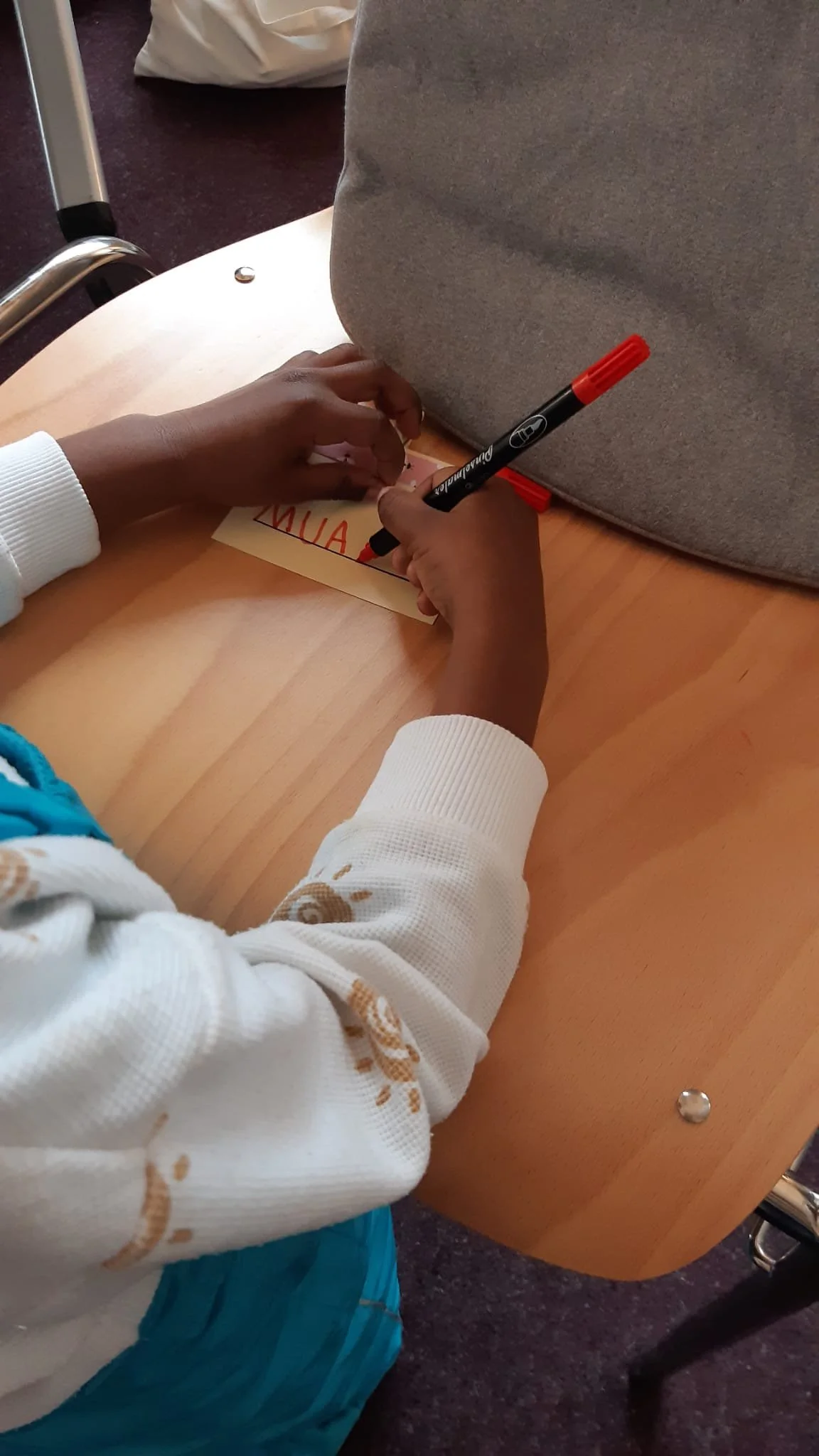 Person schreibt auf einen kleinen Zettel mit einem schwarzen Marker, auf dem "AIN" zu lesen ist, während sie an einem runden Holztisch sitzt.