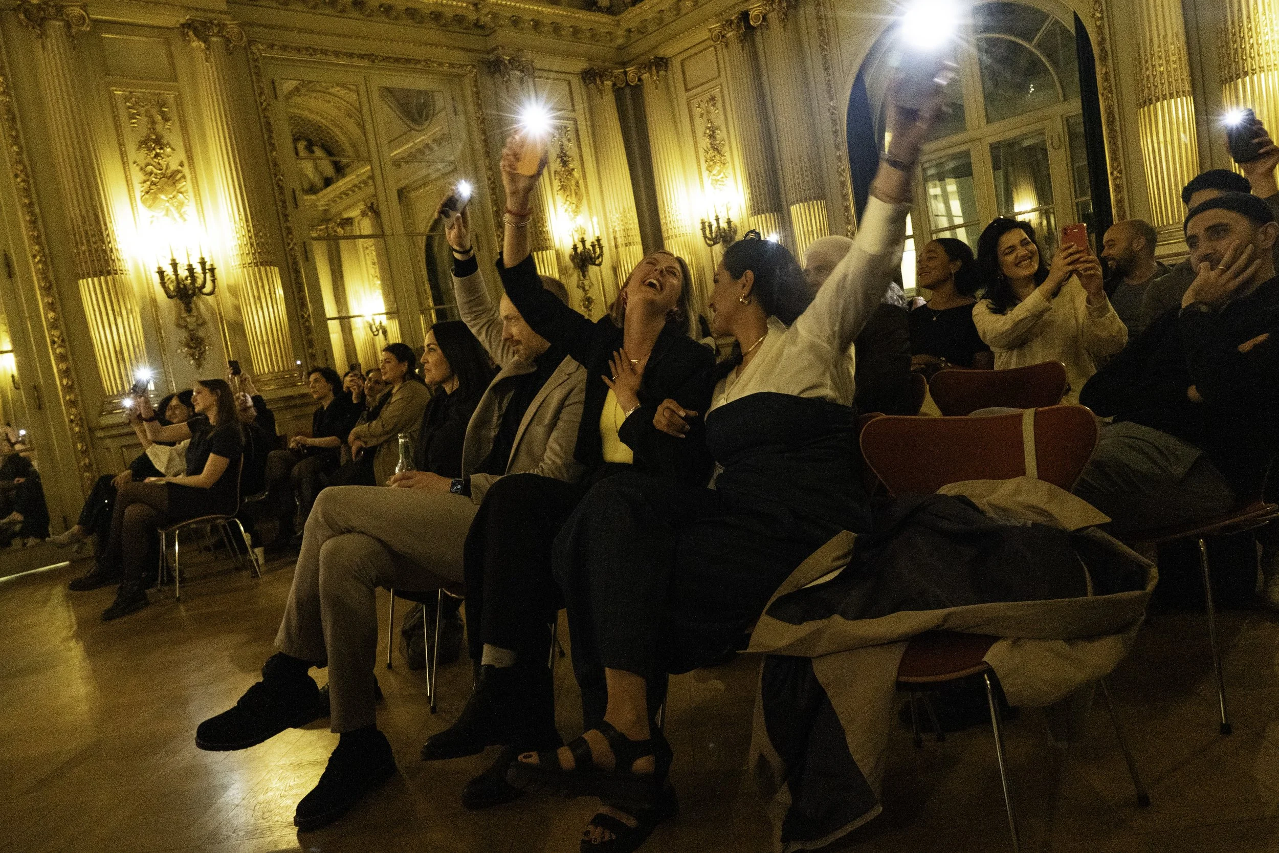Menschen in einem opulenten Saal sitzen und lachen. Einige nehmen Fotos oder Videos mit ihren Handys auf, während sie während einer Veranstaltung oder Feier in entspannter Stimmung sind.