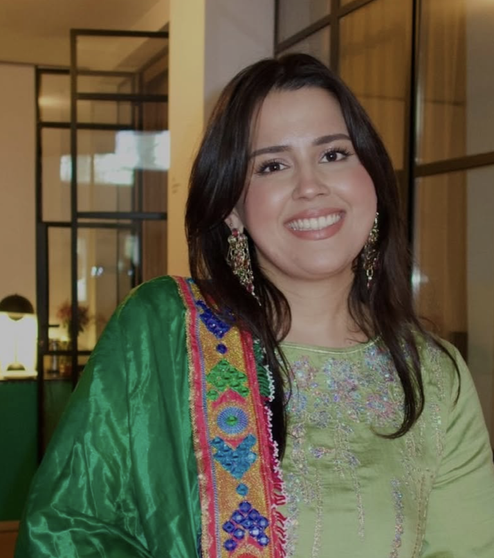 Eine lachende Frau mit dunklem, mittellangem Haar trägt traditionelle indische Kleidung und große Ohrringe, im Hintergrund ein modern eingerichteter Raum.