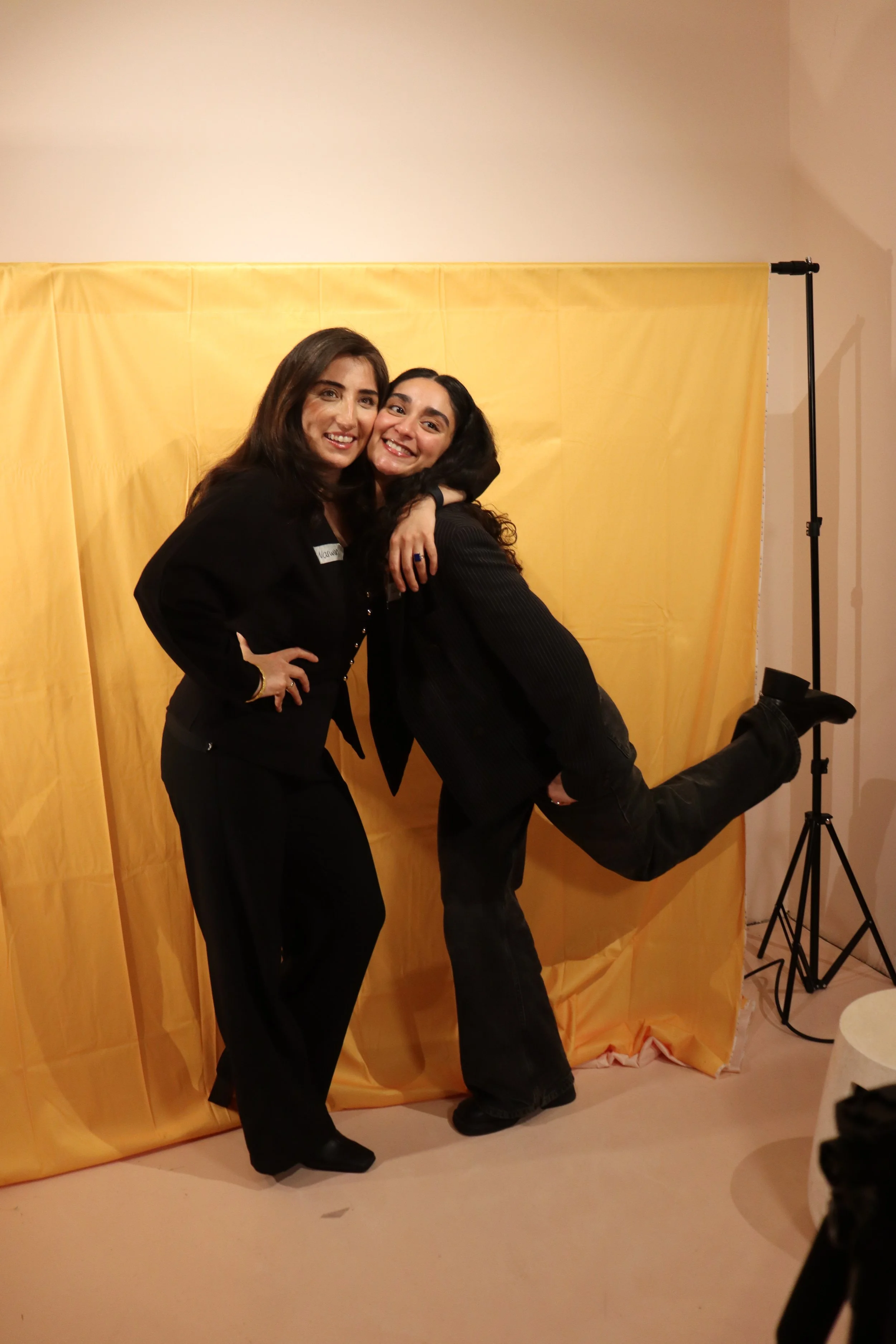 Zwei Frauen in formeller Kleidung posieren vor einem gelben Hintergrund bei einer Veranstaltung, eine lächelt direkt in die Kamera, während die andere kuschelt und ihre Fuß hochhebt.